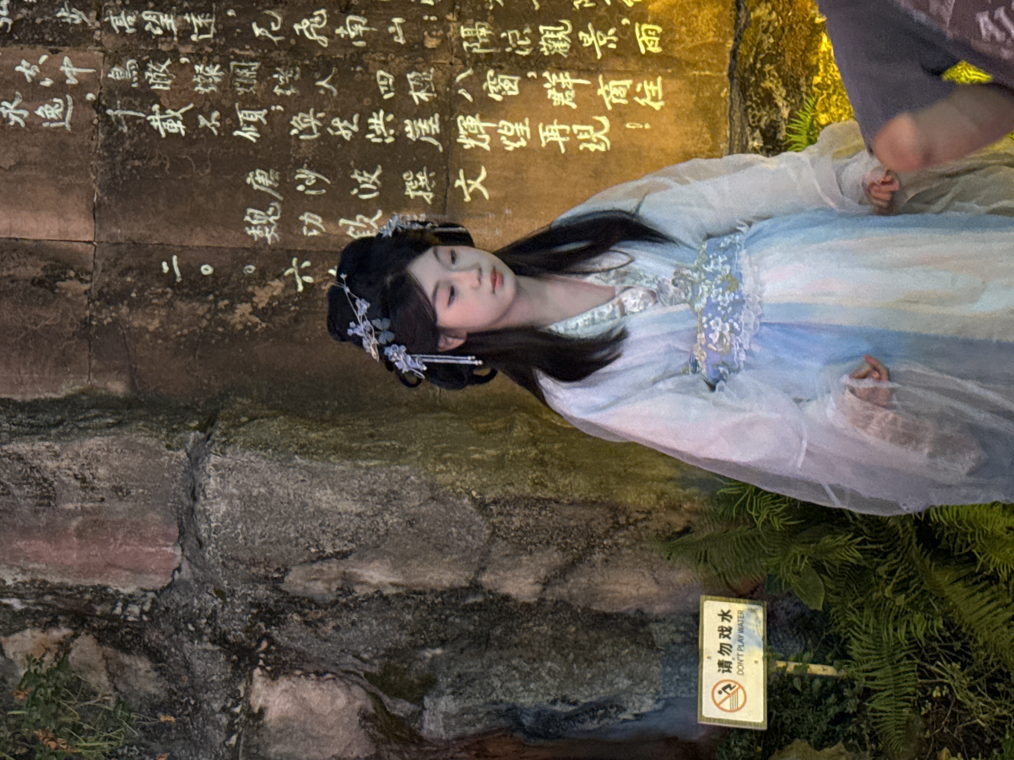 A young East Asian woman, likely in her late teens or early twenties, stands in front of a textured stone wall adorned with white Chinese calligraphy. She is dressed in a traditional Chinese Hanfu outfit, featuring a light blue and white flowing robe with intricate detailing at the waist and sleeves. Her hair is styled in an updo with a delicate floral accessory. The woman has fair skin, dark hair, and is looking downwards with a serene expression. To the left of the woman, a small sign with Chinese and English text reads "DO NOT PLAY WATER." The surrounding environment appears to be an outdoor setting, possibly a garden or a historical site, with lush green foliage visible in the lower left corner. The calligraphy on the wall suggests a cultural or historical context, possibly within the Yuzhong District of China, known for its rich heritage. The lighting in the scene is dramatic, with warm yellow and orange tones illuminating the wall and contrasting with the cooler tones of the woman's attire and the greenery. A portion of another person's arm, wearing a grey shirt, is visible in the lower right corner, suggesting the presence of onlookers.