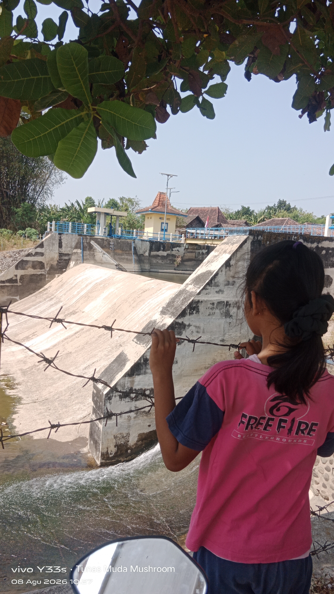 A young girl, seen from behind, stands near a concrete dam with water flowing over it. She is wearing a pink t-shirt with "FREE FIRE" printed on the back, and dark blue shorts. Barbed wire is in the foreground, and a tree with green leaves hangs overhead. In the background, there are buildings with red roofs, vegetation, and a clear blue sky. The timestamp on the bottom left indicates the image was taken on August 8, 2026, at 10:27 AM, with the device "vivo Y33s" and location "Tunas Muda Mushroom", likely referring to a place in Tanjung, Indonesia.