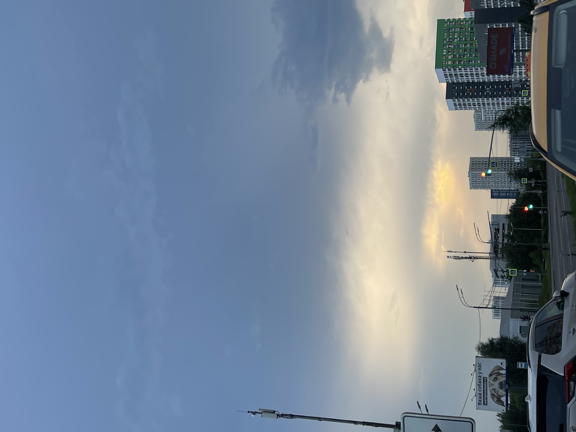 The image depicts a cloudy sky over a city street in Moscow, Russia. The sky is predominantly overcast, with patches of lighter clouds and a hint of sunset or sunrise in the distance, casting a warm glow. In the foreground, a white SUV is parked or moving along the street. Across the street, there are several tall apartment buildings, some with brightly colored facades, including a green one. A billboard advertising dog services is visible on the left, featuring an image of a dog. Traffic lights and streetlights are present, indicating an urban environment. A cell tower stands prominently on the left side of the image. Several cars are visible on the road, and a few pedestrians can be seen in the distance. The overall scene suggests a typical urban setting during twilight hours.