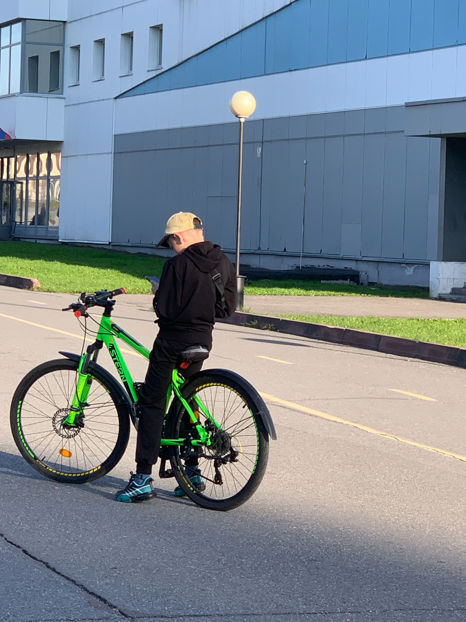 A person wearing a tan baseball cap, black hoodie, and black pants stands beside a bright green bicycle. The person is looking down at a mobile phone in their hands. The bicycle is a mountain bike with black tires and rims with yellow accents. The scene appears to be outdoors on an asphalt road with a yellow line marking. In the background, there is a modern building with a gray and blue facade, and a street lamp with a spherical white light. The ground is lined with grass in some areas. The lighting suggests it is daytime, with shadows cast on the road. The location is Prokopyevsk, Russia.