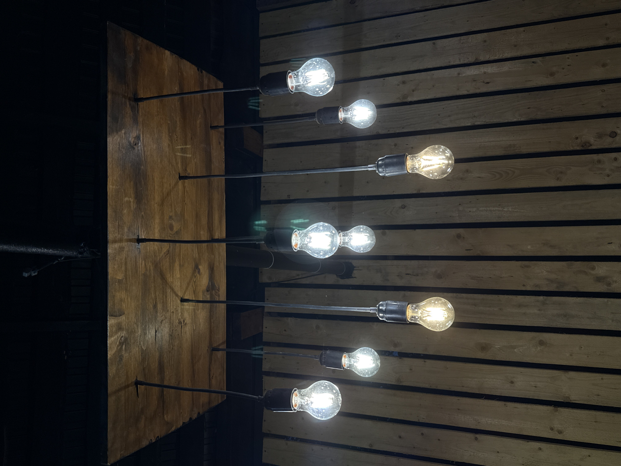 A group of eight exposed incandescent light bulbs hang from thin black cords suspended from a wooden plank ceiling. The bulbs vary in their illumination, with some appearing brighter than others. The background consists of vertical wooden slats, creating a textured, rustic aesthetic. The scene appears to be indoors, possibly in a restaurant or bar, given the style of lighting and the wooden paneling. There is no visible indication of the location being in Хохряки, Russia.
