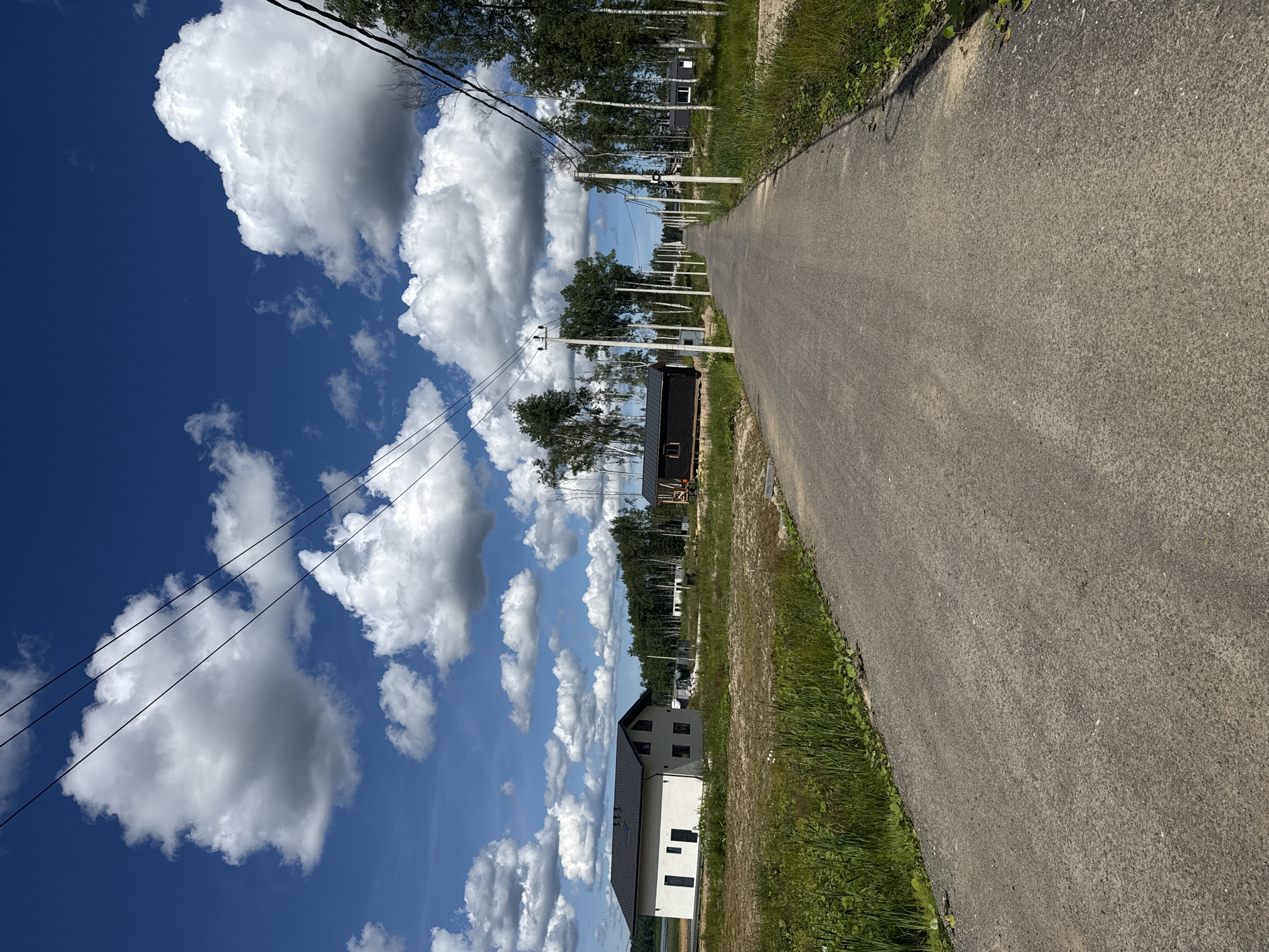 The image depicts a rural road leading through a developing residential area under a bright blue sky dotted with fluffy white clouds. The road is paved and appears to be a two-lane street. On either side of the road, there are power lines supported by white poles. Several modern houses, some appearing to be under construction, are scattered throughout the landscape. These houses are a mix of architectural styles, with some featuring white facades and dark roofs, while others are smaller, dark-colored structures. Tall, slender trees, predominantly birches, are interspersed among the houses and line the sides of the road. The ground is covered in dry grass and dirt, indicating a natural, undeveloped environment. In the distance, a body of water can be seen to the left, and a dense forest borders the area to the right. The scene suggests a quiet, suburban or rural setting, possibly a new development on the outskirts of a town. The specific location in Zashchita, Russia is not visually evident from the image itself.