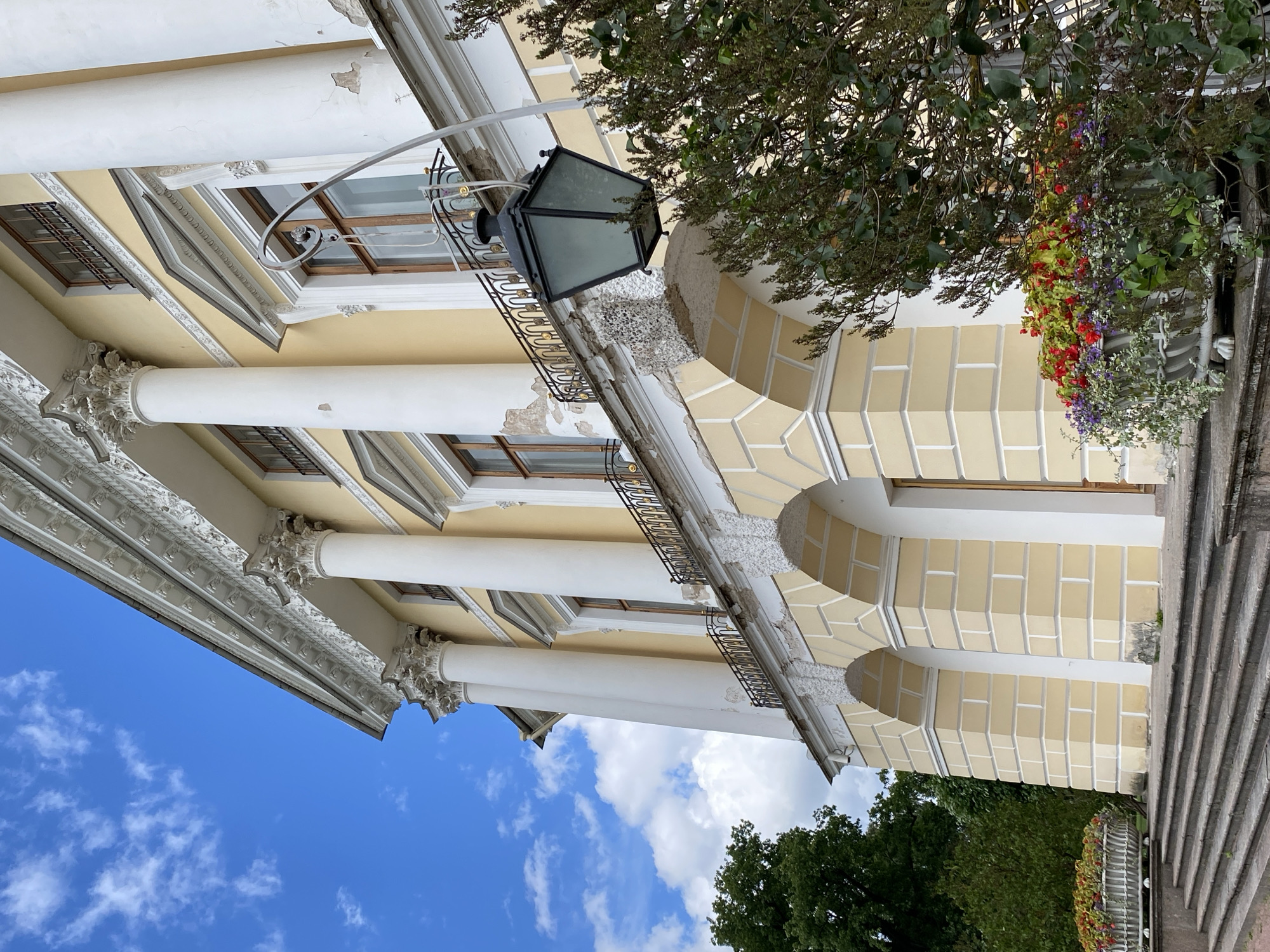 A view of the Pavlovsk Palace in Pavlovsk, Russia, with its neoclassical architecture. The palace is painted a pale yellow and features large white columns, arched entrances, and ornate decorative elements. A black lantern hangs from an ornate metal bracket on the facade. In the foreground, there are steps leading up to the palace and a planter filled with red, purple, and green flowers. Trees are visible on the left and right sides of the image, and a bright blue sky with scattered white clouds forms the background. The palace appears to be in a state of disrepair, with chipped paint and exposed concrete on some of its architectural details.