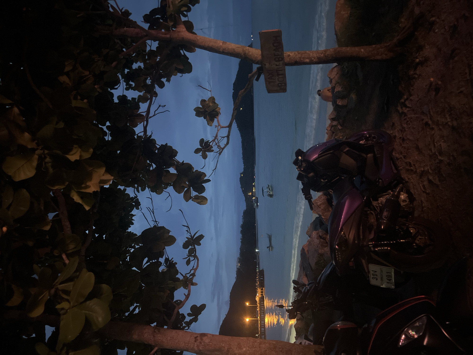 The image depicts a dimly lit scene at dusk on a beach, likely in the Ko Pha-ngan District of Thailand. In the foreground, two motorcycles are parked on the sandy shore amongst rocks. One motorcycle is predominantly purple, while the other is black with red accents. A wooden sign is affixed to a tree on the right side of the frame, appearing to read "TAXI BOTTLE BEACH." Beyond the motorcycles, the calm sea stretches to the horizon, with gentle waves lapping at the shore. A pier with lit lamps extends into the water on the left, reflecting light onto the sea. Several small boats are visible on the water, along with distant hills or mountains forming the coastline. The sky is a deep blue, transitioning from twilight to night, with hints of clouds. A lone figure sits on the rocks near the water's edge on the right side of the image. Lush green foliage from trees overhangs the scene from the top. The overall atmosphere is serene and peaceful, suggesting a quiet evening on a tropical beach.
