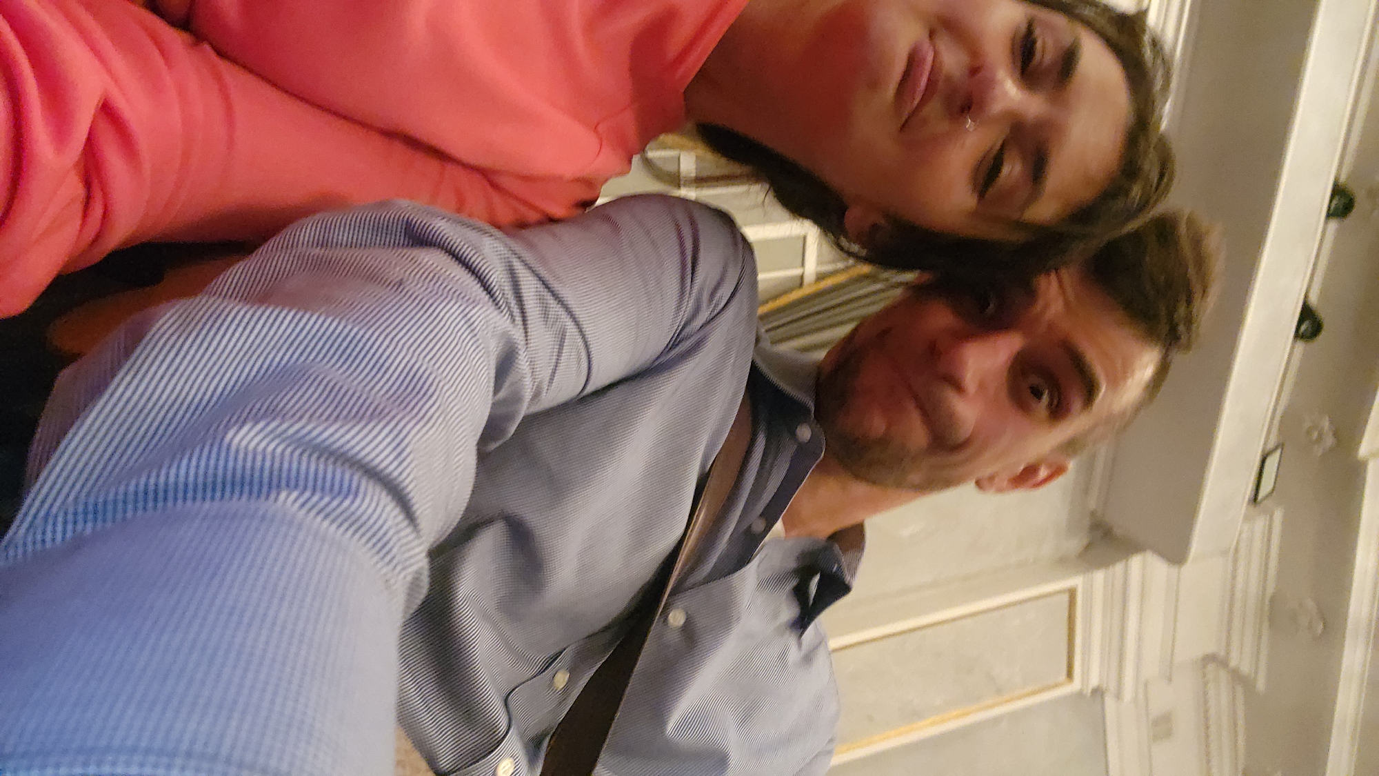 This rotated selfie captures a man and a woman indoors. The man, in the foreground to the right, is wearing a blue pinstriped button-down shirt with white buttons. He has short dark hair and light facial stubble, looking directly at the camera with a subtle smirk or smile. A dark strap, possibly from a bag, is visible over his shoulder.
Behind and to the left of him, a woman is seen wearing a coral or pink long-sleeved top. Her face is partially visible, characterized by dark hair, a nose ring, and a relaxed expression. Both individuals appear to be in good spirits.
The background features ornate white ceiling or upper wall molding with classical designs, suggesting a grand or historically styled interior. A dark, rectangular object, possibly a framed picture or sign, is visible on the wall behind the man. The lighting appears to be artificial, indicative of an indoor setting. The image is taken from a low angle and is rotated approximately 90 degrees clockwise, giving it a dynamic and candid feel. No discernible text is visible in the image. Given the location context, this scene is set in Moscow, Russia, likely within a building with classical architectural elements.