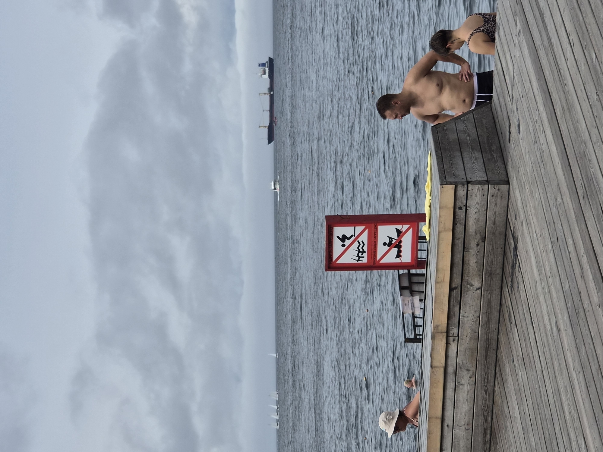 In Vladivostok, Russia, on an overcast day, a scene unfolds by the sea. In the foreground, a wooden pier stretches into view, its weathered planks laid out in parallel. To the left, a woman wearing a sun hat and swimsuit is partially submerged in the water, her body angled away from the camera. Further in the water, another person is visible.
Dominating the middle ground is a red and white sign depicting forbidden activities: jumping into the water and paddling a canoe. Adjacent to this sign, a large cargo ship sails on the horizon, dwarfed by the expanse of grey sky. In the distance, several sailboats with white sails are scattered across the choppy water.
On the right side of the pier, a shirtless man stands with his hands on his hips, looking out at the sea. Beside him, a woman wearing a leopard-print swimsuit looks down. The water is a deep, muted blue, reflecting the overcast sky.