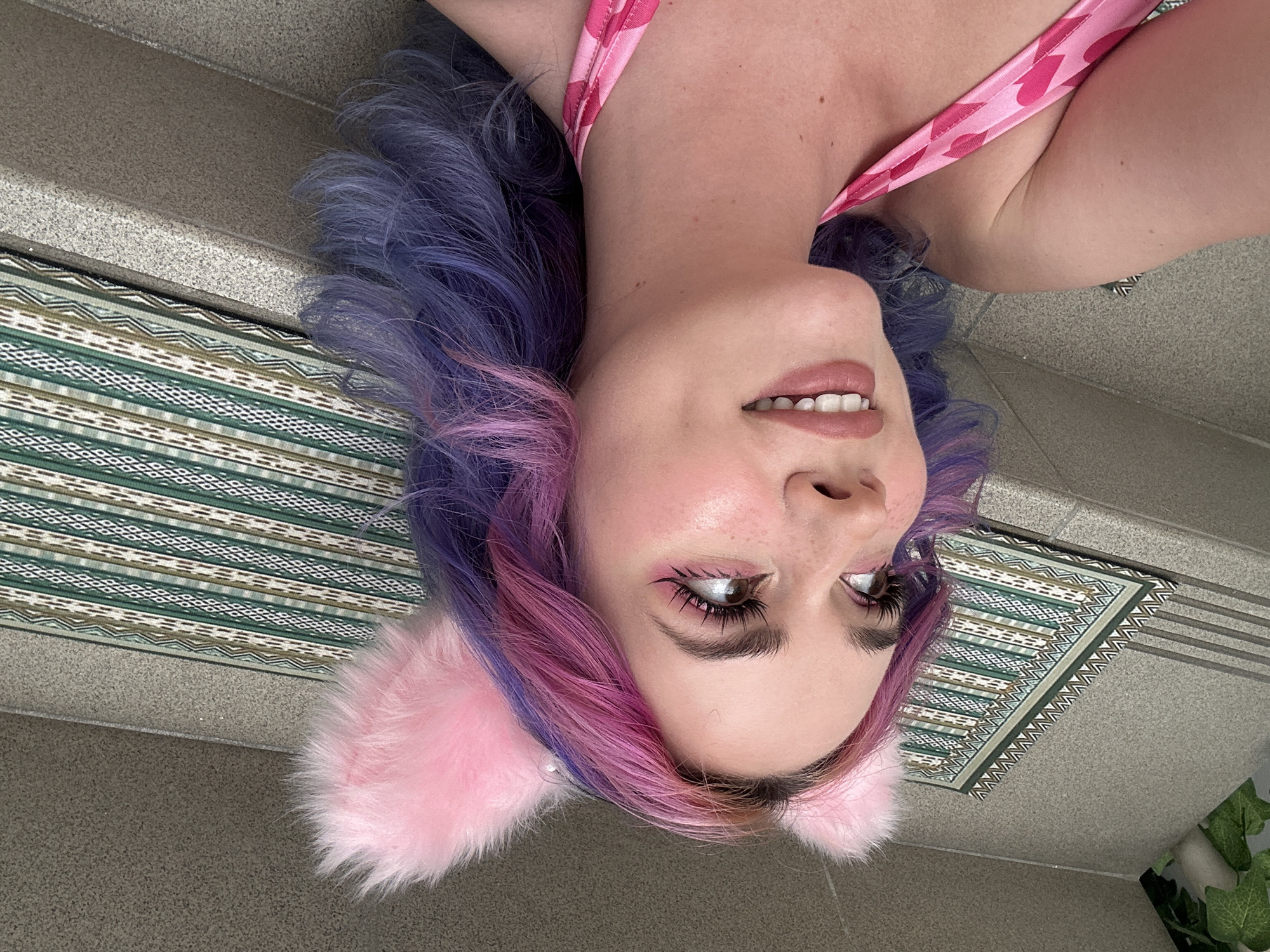 A woman with pink and blue hair, wearing pink cat ears and a pink top, lies on a gray tiled surface with a green and brown patterned rug. The setting appears to be an urban outdoor area. There is no definitive information in the image to specify the exact location as the City of Zagreb, Croatia.