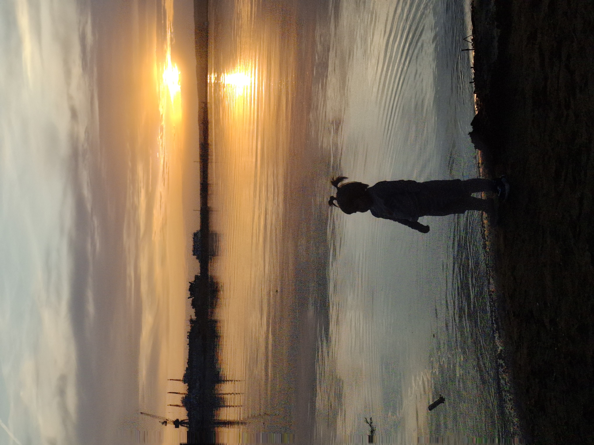 A sunset silhouettes a child at the edge of the water, presumably the Kama River, at sunset in the city of Chaikovsky, Russia. The child stands near the edge of the water, likely a bay or inlet, the setting sun casts an orange and golden hue across the water and sky, while a cityscape with a crane is visible in the distance. The foreground is dark, obscuring details except for the outline of the child.