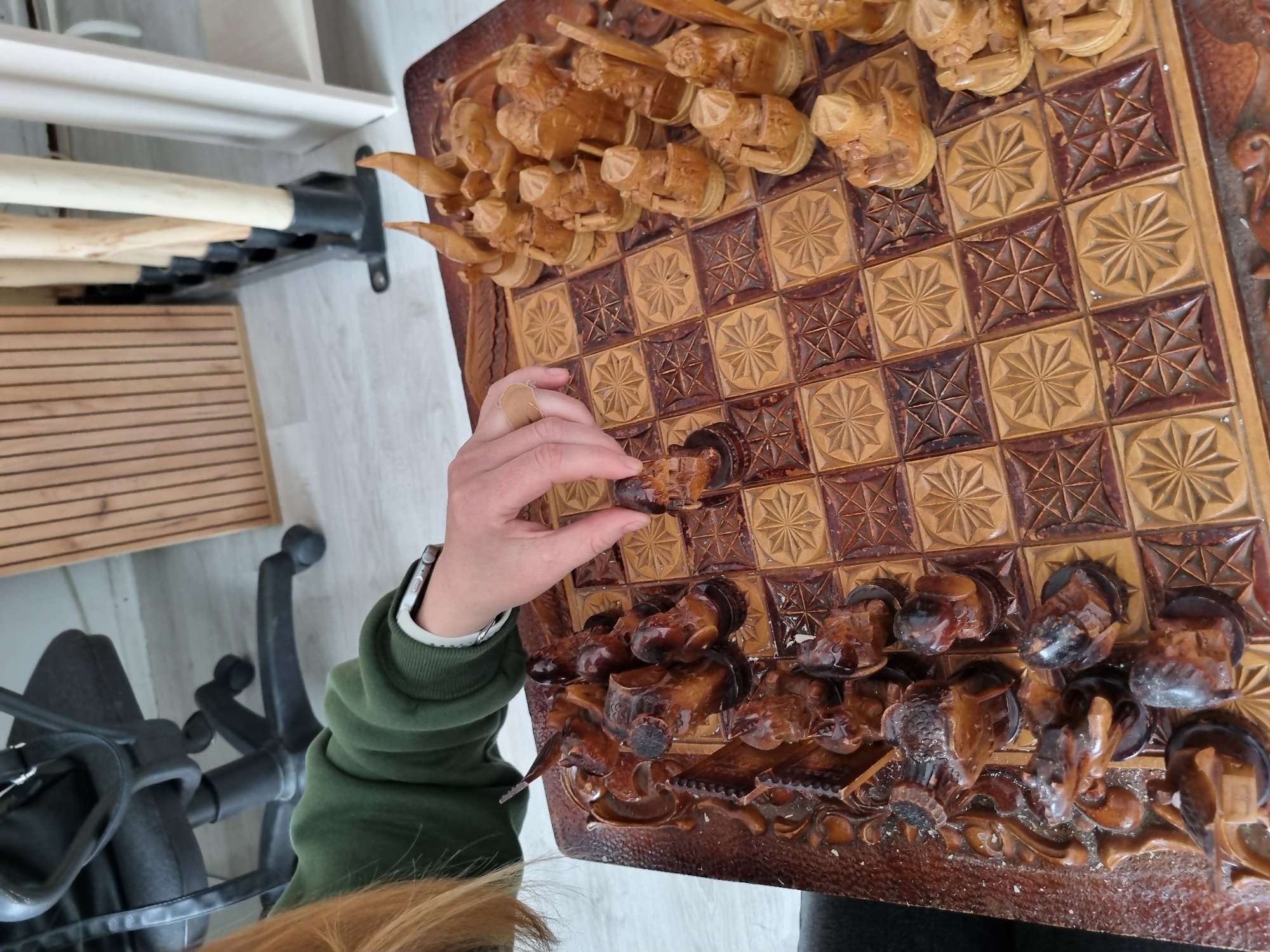 A person is holding a dark-colored chess piece above a carved wooden chess board. The chess board is brown and tan and features highly detailed, carved chess pieces along the edges. On the left side of the image, there is a gray floor, a black office chair, and a wooden rack against a white wall. There is no evidence to indicate this image was taken in Chaikovsky, Russia.