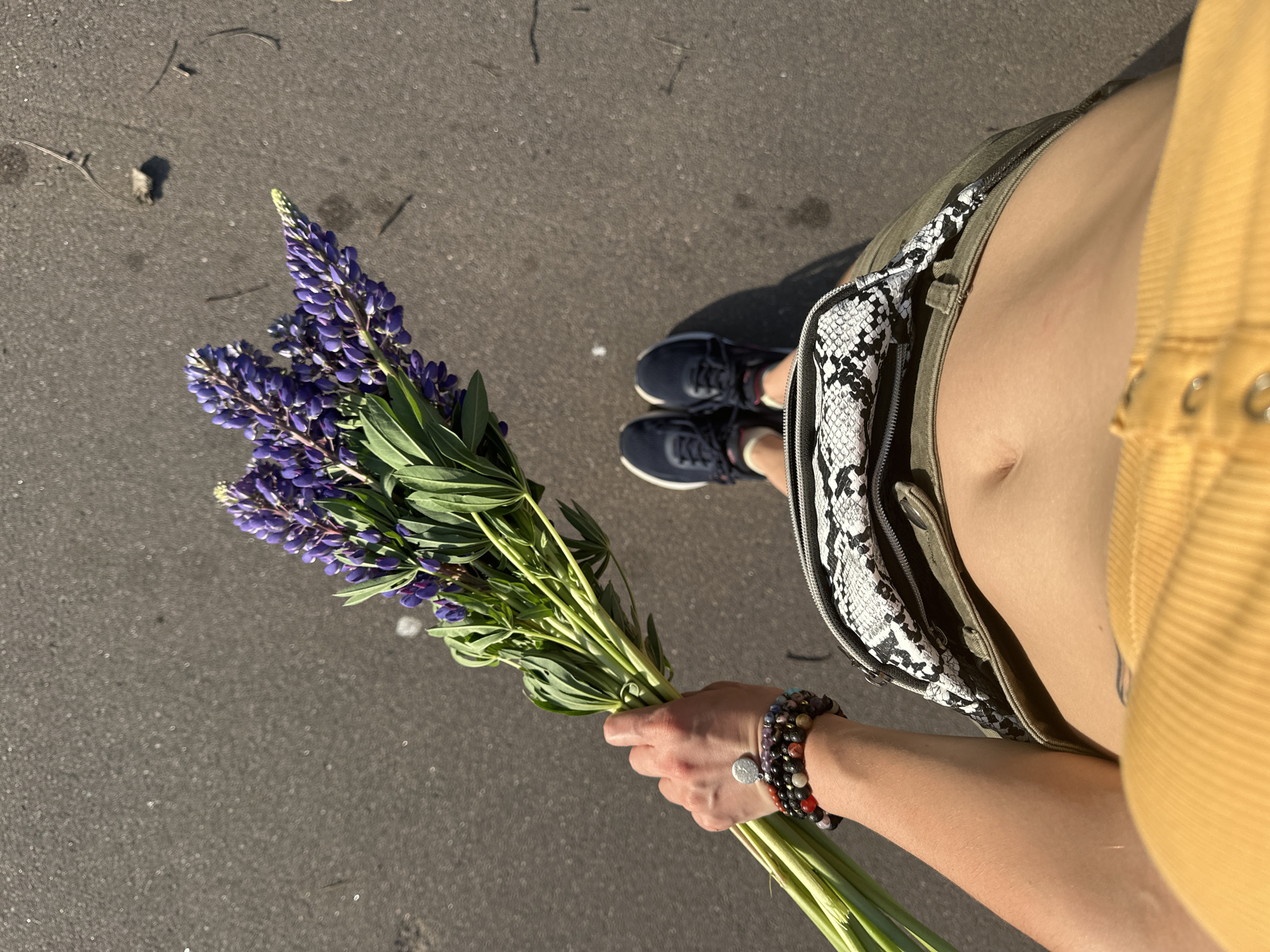 A first-person perspective shot from above shows a person standing on a paved surface, holding a bouquet of purple lupine flowers. The person is wearing a yellow top, a green skirt with a snake-print fanny pack, and dark sneakers. The pavement is dark gray with some visible debris. The image does not provide information about the specific location in Vladimir, Russia, or any interactions or actions beyond the static pose.