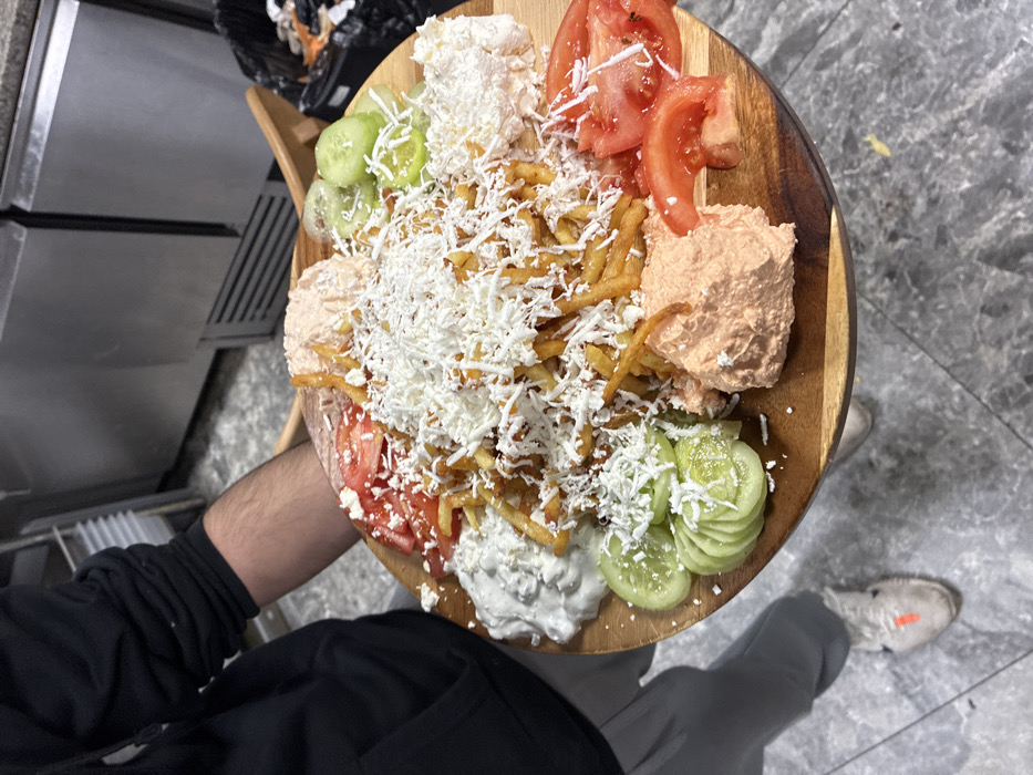 A person, wearing a black long-sleeved top and grey pants with white sneakers, holds a large round wooden platter laden with food in an indoor, commercial kitchen or food service environment in Sofia, Bulgaria. The central focus of the platter is a substantial mound of golden-brown french fries heavily garnished with shredded white cheese. Surrounding the fries, arranged in distinct segments, are various accompaniments: multiple slices of fresh red tomatoes, several slices of green cucumbers, a portion of a white creamy dip (resembling tzatziki), a portion of an orange-pink creamy dip, another portion of a white, chunky spread, and a second portion of the orange-pink creamy dip. The background reveals stainless steel refrigerators or counters and a grey tiled floor. A black waste bin is partially visible on the far right. The person's arm, lower torso, and legs are visible, presenting the platter directly towards the viewer.