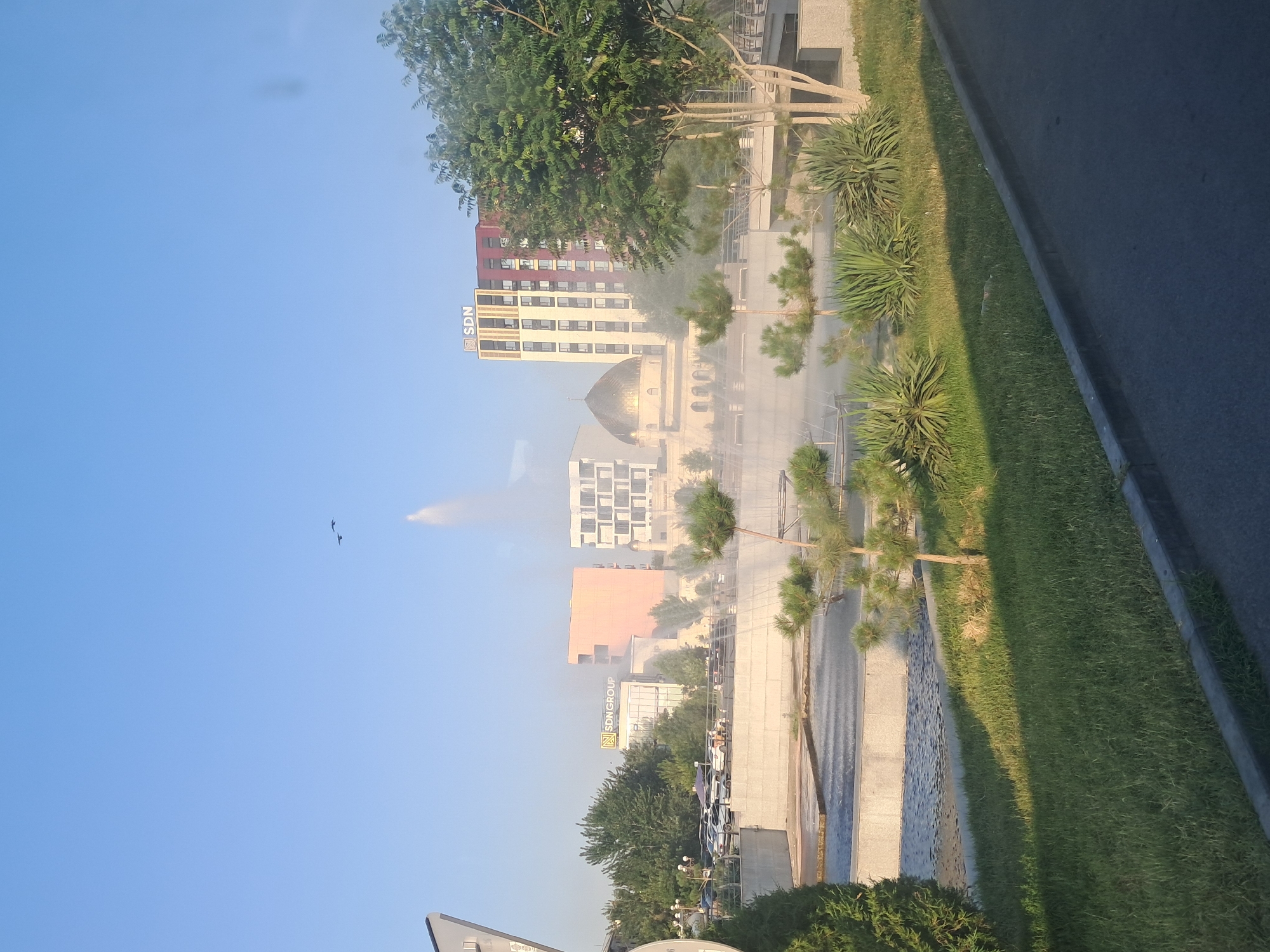This image depicts a street scene in Shymkent, Kazakhstan, with a focus on modern architecture and landscaping. In the background, several buildings are visible, including a prominent multi-story building with a red facade and a beige lower section featuring a "SDN" logo. Adjacent to it is a building with a golden dome, possibly a mosque, and another building with a grid-like facade. The foreground features a paved walkway, a grassy area with manicured bushes and trees, and a road. The presence of mist or fog suggests a humid or possibly artificial misting system is in operation, creating a diffused atmosphere. The lighting indicates it is daytime, likely morning or late afternoon due to the angle of the shadows and the soft light. The sky is clear and blue, with two birds in flight visible in the upper left portion of the frame. The overall impression is one of urban development with an emphasis on public spaces and greenery. No people are clearly discernible in the image, but the presence of parked cars suggests some activity. The "SDN" text is clearly visible on one of the buildings.