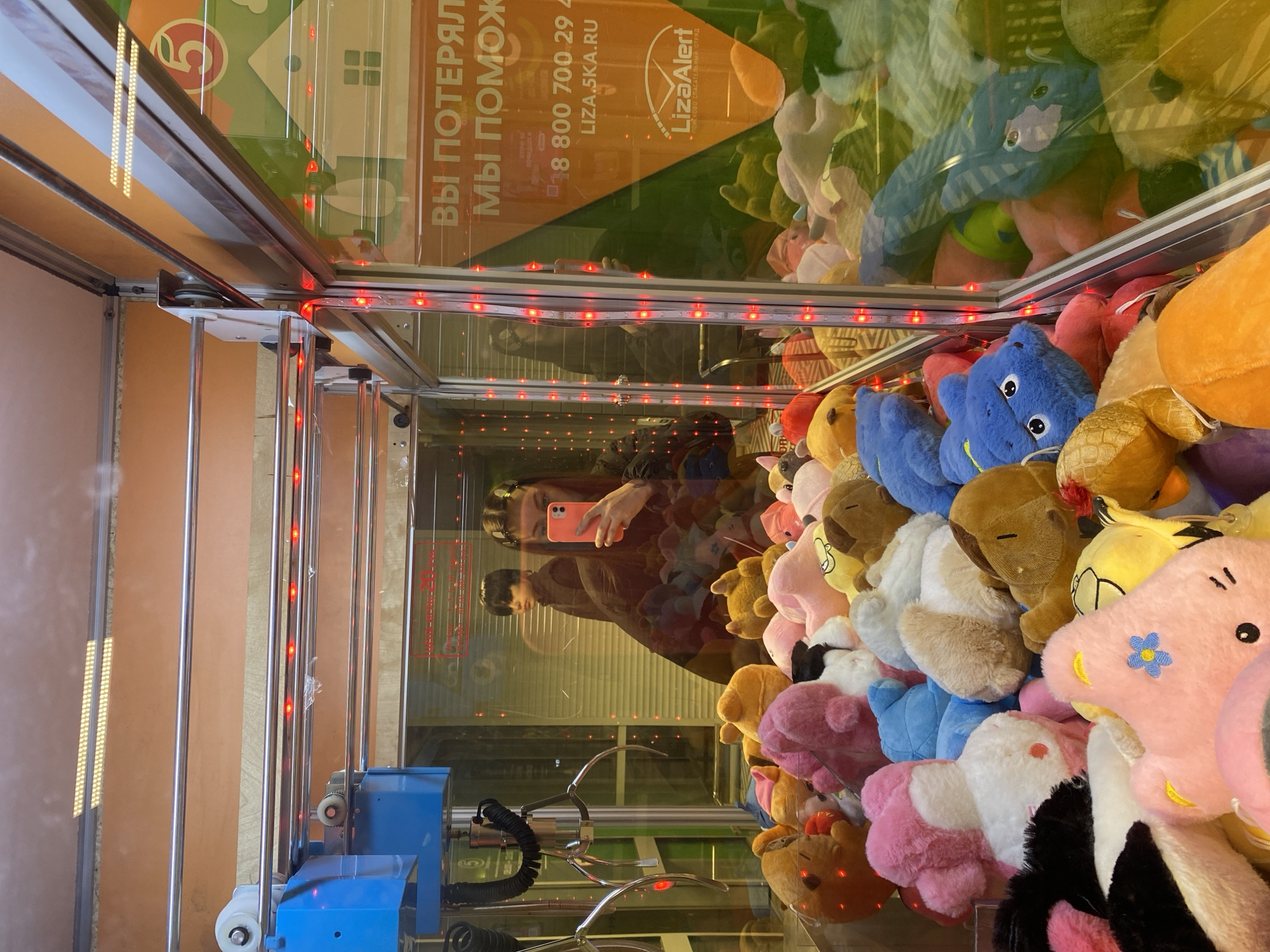 The image depicts the inside of a claw machine, presumably in Votkinsk, Russia. The machine is filled with stuffed animals of various colors and shapes. A person, likely taking the photo, is reflected in the glass, holding a pink smartphone. The machine's claw is visible, positioned among the toys. Red LED lights illuminate the interior of the machine. A sign with Russian text and the LizaAlert logo is visible on the machine's side. The environment surrounding the machine appears to be an indoor space, with some architectural details visible in the reflection.