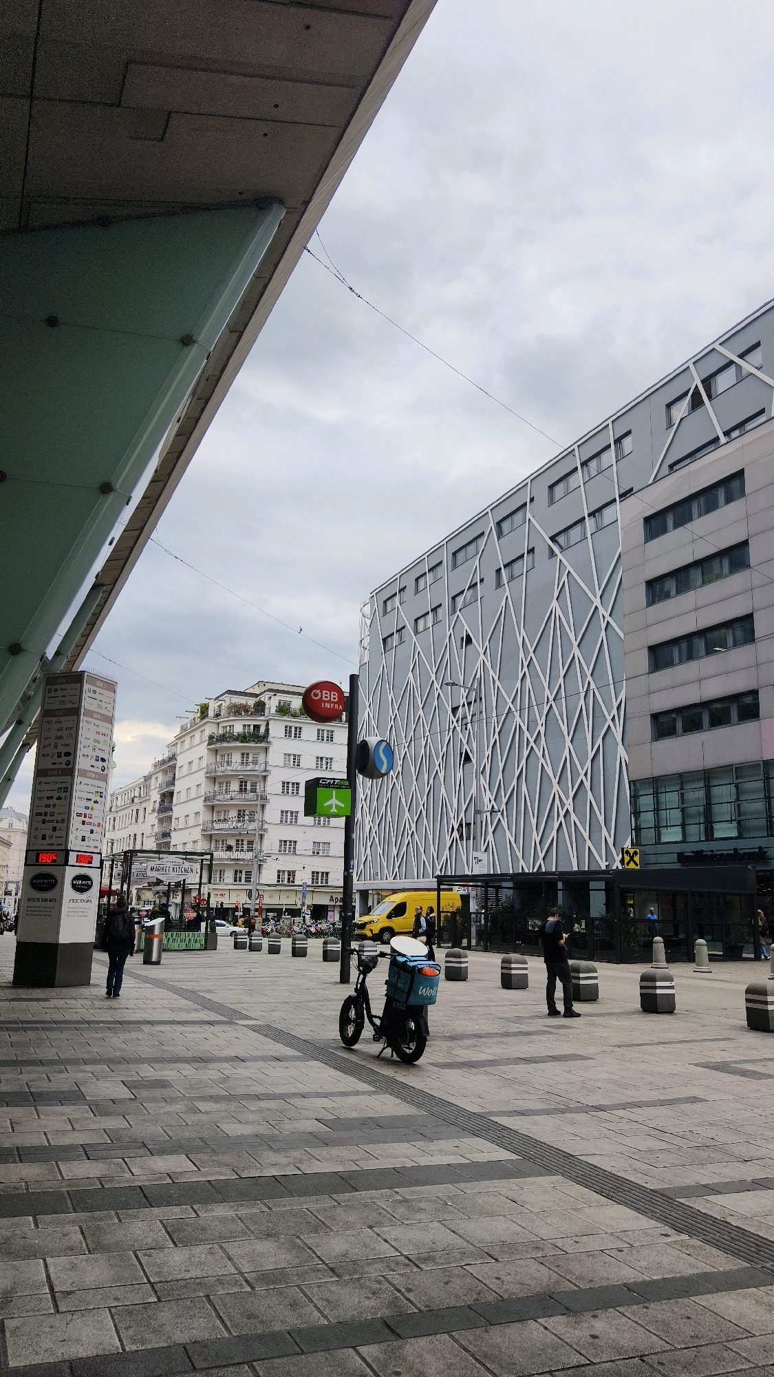 The image shows a street scene in Vienna, Austria, likely near a transportation hub or a busy commercial area. In the foreground, a modern building with a glass and concrete overhang shades a paved pedestrian area. The pavement is made of large, light grey rectangular tiles with darker grey borders. To the left, a tall, rectangular pillar displays various advertisements and logos, including "1906" and "MARKET KITCHEN". A person is walking away from the pillar towards the left.
In the mid-ground, a black electric scooter with a blue delivery box is parked on the pedestrian walkway. The box has the "Wolt" logo on it. Two bollards are positioned to the right of the scooter. Beyond the scooter, a bright yellow van is visible, and several people are walking or standing.
The background features several buildings. To the left, a row of white buildings with balconies and many windows is visible. In the center, a large grey building with a distinctive criss-cross white pattern on its facade dominates the scene. A red circular sign with "OBB INFRA" is attached to a pole in front of this building. A green rectangular sign with an airplane icon is also present on a pole. The sky is overcast and grey. The scene depicts a typical urban environment with commercial activity and pedestrian movement.
