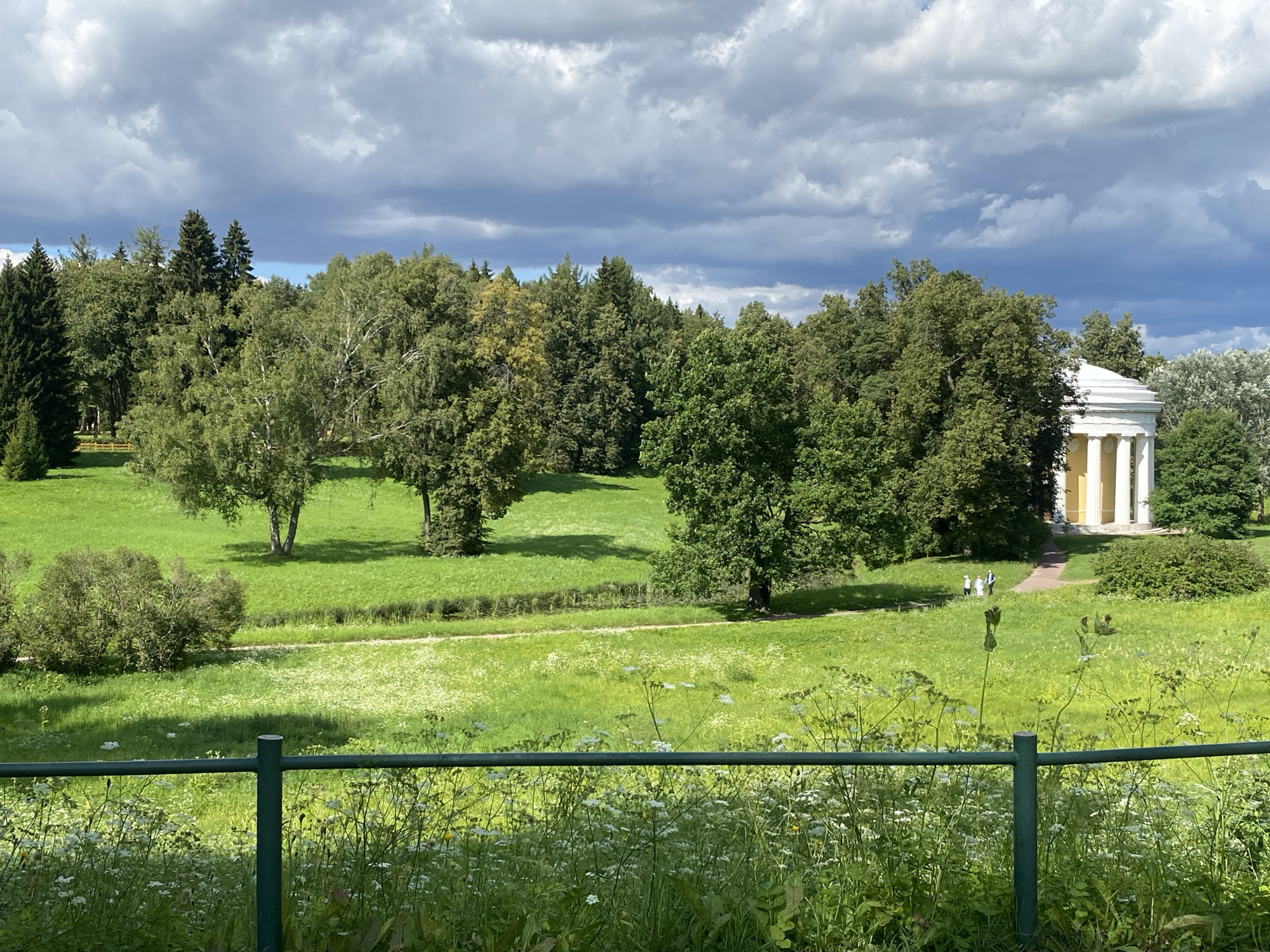 The image depicts a vast green landscape under a dynamic cloudy sky. In the background, a white classical building with columns is visible, surrounded by lush green trees. Several people are walking on a path leading to the building. The foreground features a green metal fence and a field of white wildflowers. The scene appears to be set in a park or garden. This image was taken in Pavlovsk, Russia.