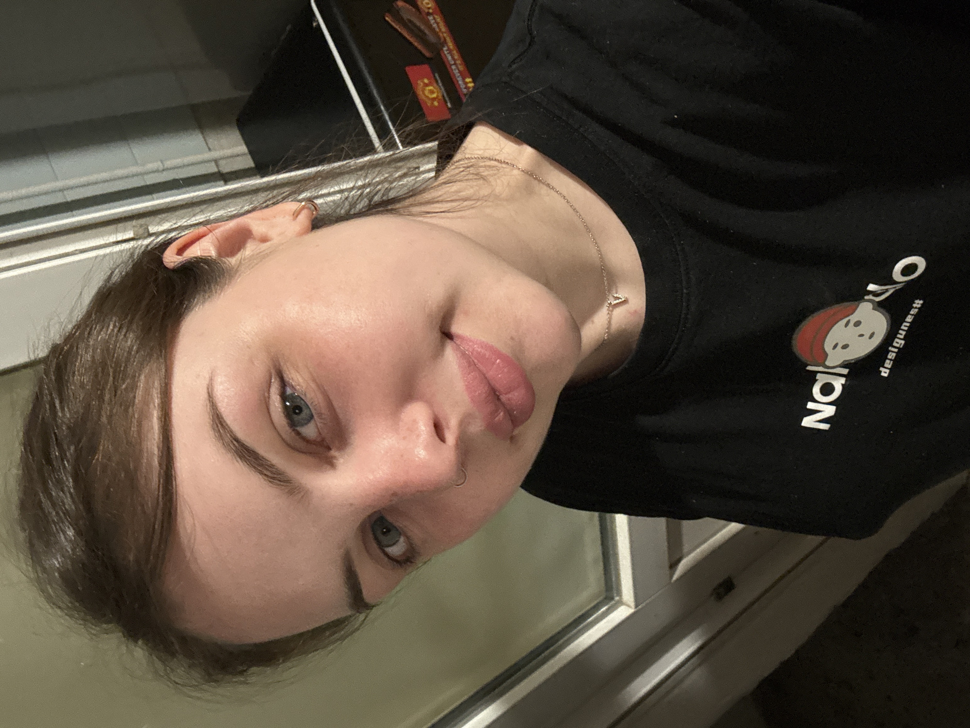 A close-up, angled headshot of a fair-skinned woman with light blue eyes. She has brown hair, a pierced nose, and is wearing a necklace. She appears to be inside, with a window frame visible behind her. She is wearing a black t-shirt with a white logo that includes a graphic and the word "designsnest". The location is not discernible.