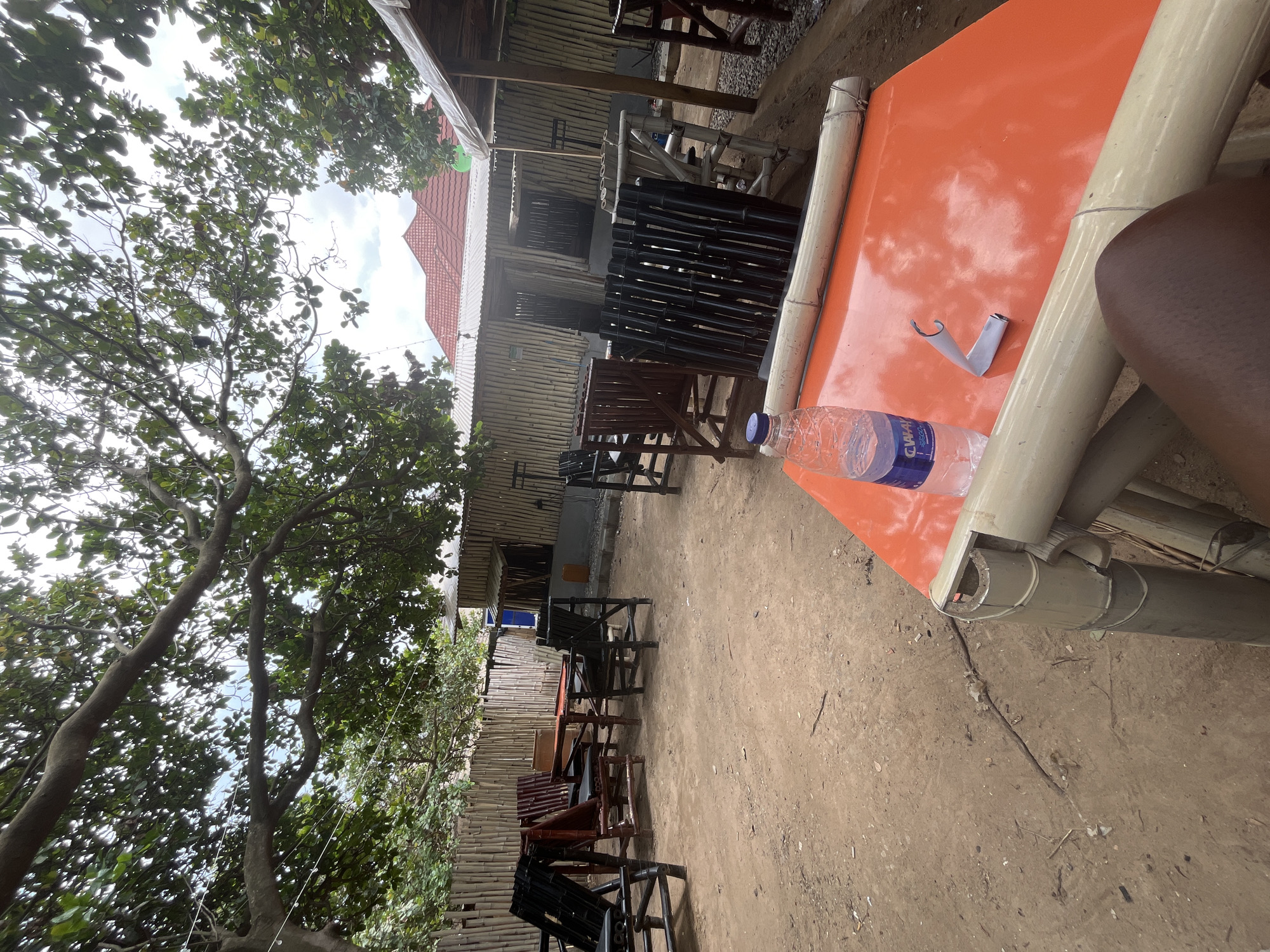 The image depicts an outdoor seating area, likely a restaurant or cafe, in Abuja, Nigeria. The scene is oriented vertically, with the top of the image showing trees and the sky, and the bottom showing the ground and furniture.
The setting appears to be rustic and casual, with many elements made of bamboo, including the structure of the building in the background, the fencing along the side, and the legs of the table in the foreground. The building has a tiled roof and bamboo walls. Various chairs, made of dark wood and bamboo, are arranged in rows, suggesting that this is a place for dining or relaxation. Some chairs are empty, while others are partially obscured.
On an orange table in the foreground, a plastic water bottle with a blue and white label is visible. The label has text that is partially visible, including "CHAMP" and "WATER". A small piece of white paper, folded in a way that suggests it might have been used to seal the bottle or as a napkin, is also on the table. A person's leg and arm, appearing to be dark-skinned, are visible at the right edge of the frame, suggesting a person is seated at the table.
The ground is covered in dirt and small stones. The lighting suggests it is daytime, possibly late morning or early afternoon, with bright, diffused light filtering through the leaves of the trees. The weather appears to be overcast or partly cloudy, as the sky is bright but not intensely sunny. The overall impression is one of a relaxed, open-air environment, perhaps a getaway spot in a tropical or sub-tropical climate.