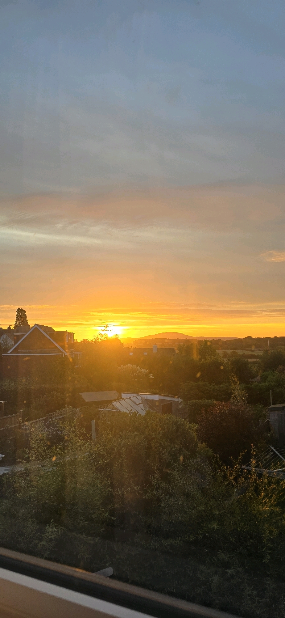 A vertical, eye-level, outdoor shot captures a vibrant sunset over Bayston Hill, United Kingdom. The sky is a dramatic gradient of warm colors, transitioning from a pale blue at the top to a brilliant orange and yellow near the horizon, suggesting the sun is low. Wispy clouds are visible throughout the sky.
The foreground is dominated by lush green foliage, with sunlight filtering through the leaves, creating a dappled effect and lens flare. The leaves are detailed, showing texture and the movement of gentle light.
In the middle ground, several houses and outbuildings are visible, nestled amongst trees and gardens. One house in particular, with a reddish-brown brick facade and a pitched roof, is prominent on the left. Other structures include greenhouses and smaller sheds. The overall impression is of a residential area with gardens and greenery.
In the background, a distant hillside is silhouetted against the bright sunset. The landscape appears to be a mix of rural and suburban, typical of the outskirts of a town or city. The lighting is soft and warm, characteristic of sunset or sunrise. The overall mood is serene and picturesque, emphasizing the beauty of the natural light and the suburban landscape.