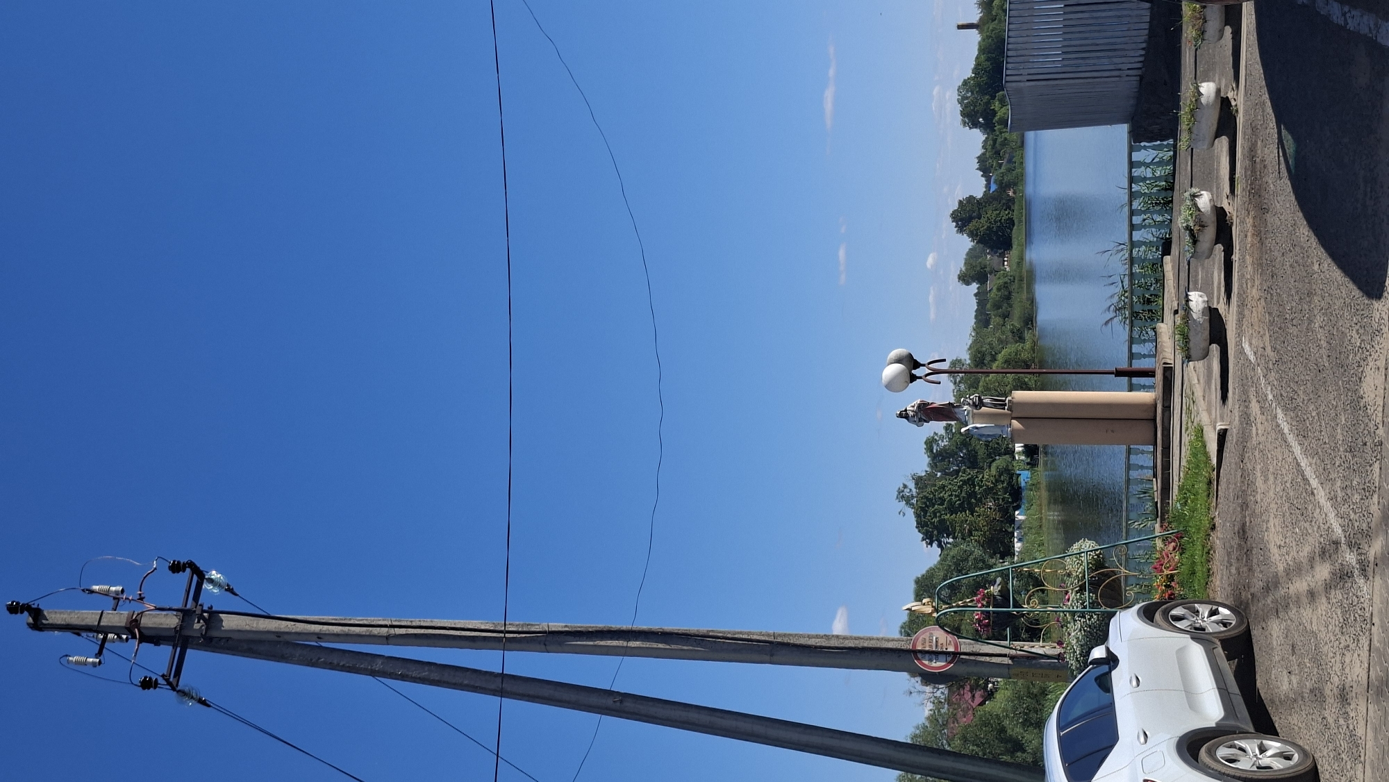 A white car is parked on the right side of the frame, facing away from the camera. In front of the car, there are several concrete planters with plants. Behind the planters, there is a paved area leading to a lake. On the left side of the frame, there is a tall telephone pole with wires. In the middle of the scene, there is a statue of a religious figure on a pedestal, with two lamps next to it. The lake is calm and reflects the clear blue sky. Trees and houses are visible on the opposite shore of the lake. The overall scene depicts a quiet, sunny day in a residential area, possibly a park or a lakeside promenade.