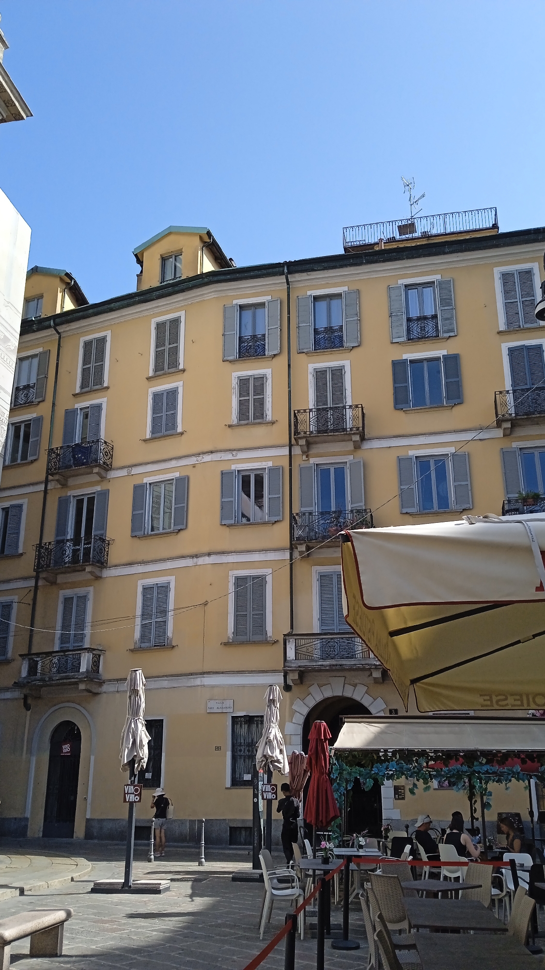 The image depicts a street scene in Milan, Italy, with a focus on the exterior of a yellow building featuring multiple windows and balconies. The building appears to be multi-story, with a clear blue sky above. In the foreground, an outdoor seating area for a cafe or restaurant is visible, complete with tables, chairs, and umbrellas. Some people are seated at the tables, suggesting a relatively casual daytime setting. The street surface is paved with cobblestones or patterned tiles. The architecture suggests a European urban environment.