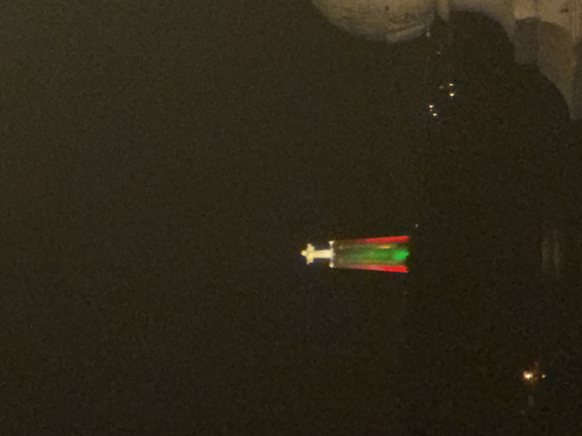 The image displays the Santuário de Cristo Rei, a monument in Almada, Portugal, overlooking Lisbon. The monument, a statue of Jesus Christ with outstretched arms, is illuminated at night. The base of the monument is lit with vertical bands of red and green light, suggesting a national flag motif or holiday illumination. A small, bright white cross is visible atop the statue. The background is dark, indicating nighttime, with distant lights from the city of Lisbon visible on the horizon. A blurred, light-colored architectural element, possibly a balustrade or part of a building, is visible in the foreground on the right side of the image. The overall scene is a nighttime view of a prominent landmark in the Lisbon metropolitan area.