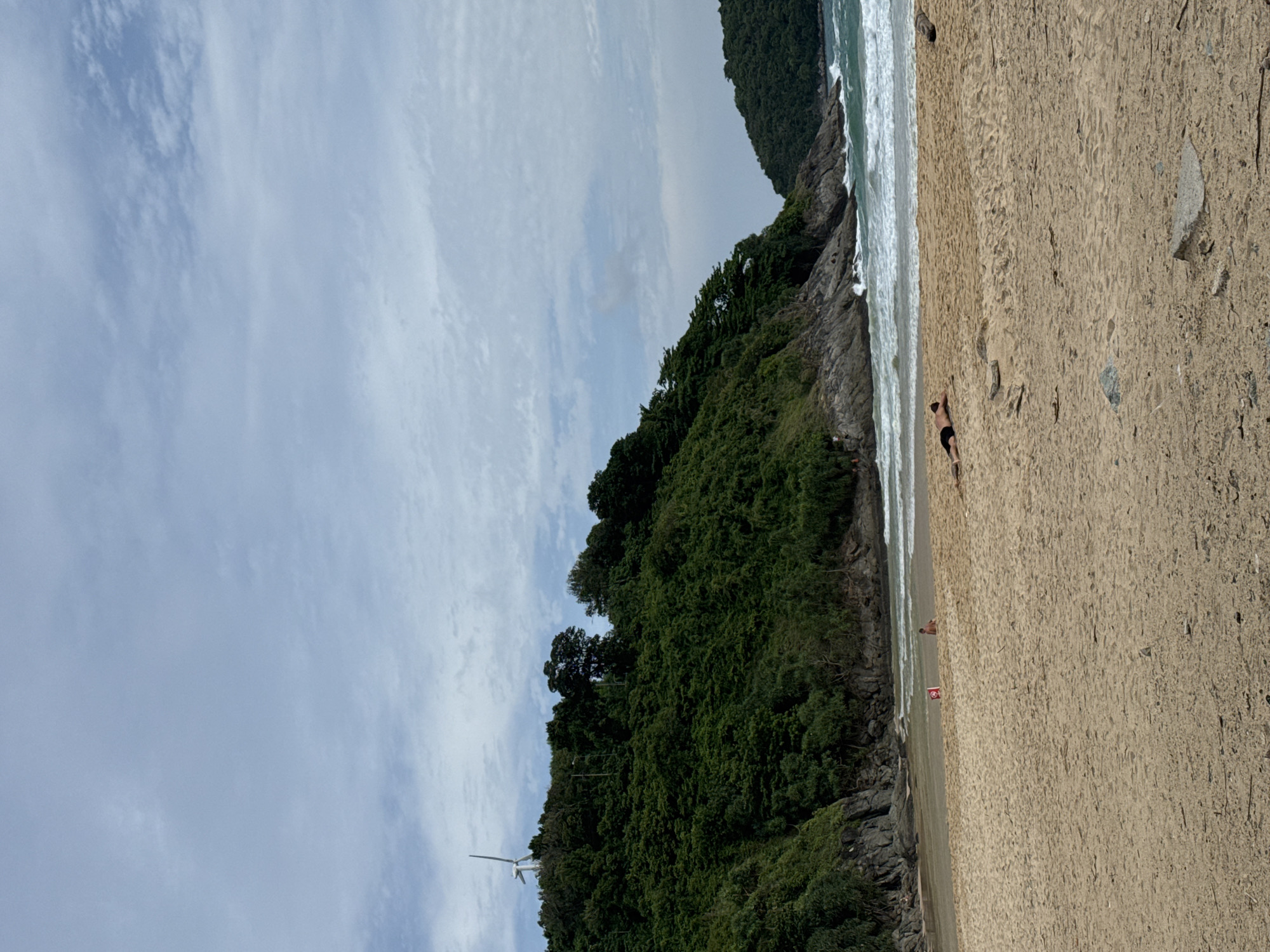 An outdoor, wide shot captures a sandy beach in Rawai, Thailand, under a cloudy sky. The foreground is dominated by a wide expanse of light brown sand with scattered debris. In the middle ground, a lone individual lies face down on the sand, wearing black shorts. Further back, near the water's edge, two other individuals are visible, one sitting and one standing. A red and white sign is placed in the sand between them. The background features a rocky coastline with lush green trees, a prominent hill with a windmill on top, and the turquoise ocean with white waves crashing onto the shore. The overall scene suggests a relatively calm and natural beach environment.