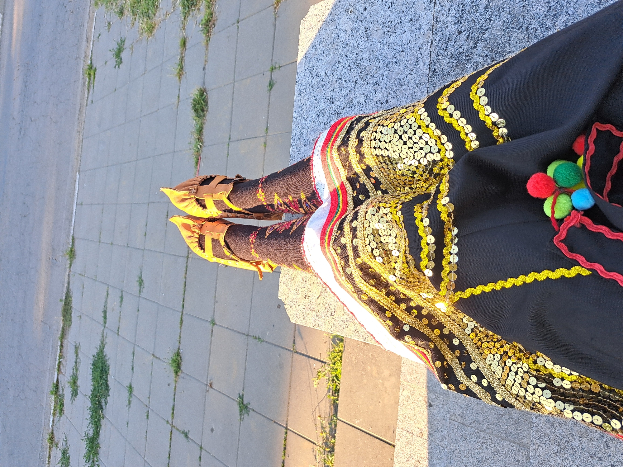 A low-angle shot shows a person's legs and feet, clad in traditional attire, as they sit on a stone ledge. The person's legs are crossed, and they are wearing dark-colored tights with embroidered patterns in red, yellow, and white. On their feet are pointed, open-toed sandals with brown leather straps.
The garment is primarily black, adorned with abundant gold sequins arranged in decorative patterns along the hem and down the legs. Bands of red, white, and yellow trim add further embellishment. Colorful pom-poms in red, green, and blue, attached by red cords, are visible on the fabric near the waist.
The setting appears to be outdoors, with a gray tiled sidewalk or pathway in the background. Patches of green grass are growing between the tiles. The sunlight casts a warm glow, suggesting it is daytime, likely late afternoon due to the long shadows. The overall mood is calm and relaxed. There is no visible text in the image.
