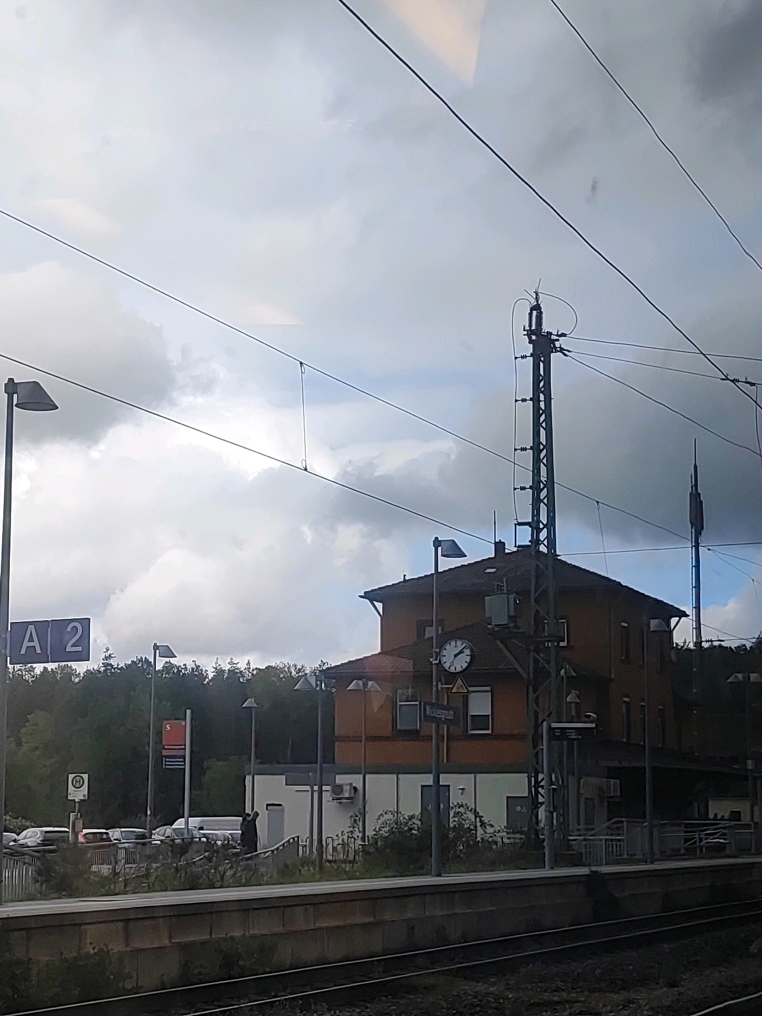 The image captures a view from a train platform in Petersaurach, Germany. In the foreground, train tracks are visible, and the platform edge with concrete and stone structures extends across the bottom. Behind the platform, a red-brick building that appears to be a train station is situated. A clock is mounted on the building's facade, showing approximately 12:35. A tall metal pylon with electrical equipment stands in front of the building, supporting overhead power lines that crisscross the sky. To the left of the station building, a sign displays "A 2" and a lamppost is present. Further left, another sign with an "H" in a circle is visible, along with parked cars. The background is filled with dense green trees. The sky is overcast with dramatic clouds. A person is seen walking on the platform near the station entrance.
