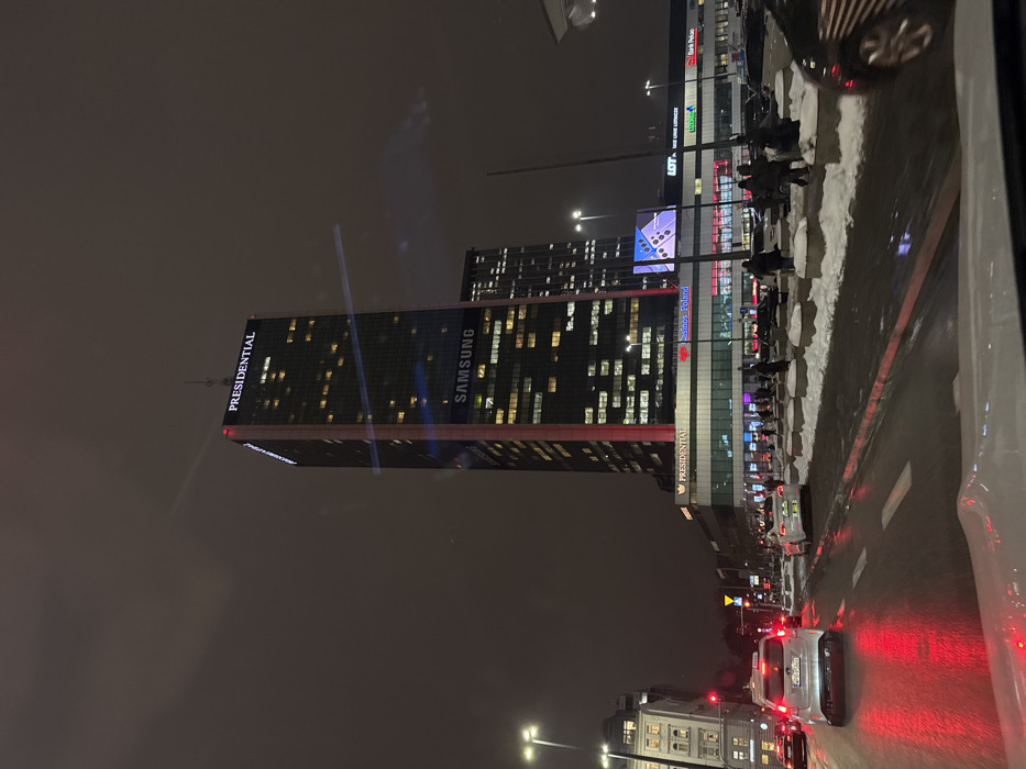 The image depicts a nighttime urban scene in Warsaw, Poland, focused on the Warsaw Trade Tower located at ul. Chłodna 51. The prominent skyscraper, a tall, dark glass structure, is illuminated with interior lights in many of its windows, red vertical light strips along its sides, and external signage displaying "PRESIDENTIAL" at its top and "SAMSUNG" mid-way down its facade.
At the base of the tower, a commercial complex features illuminated signs for businesses, including "Casinos Poland," "LOT Polish Airlines," and "Bank Pekao," among others.
In the foreground, a wet asphalt street reflects red light from vehicle taillights and a traffic signal. A white Skoda Octavia taxi with a roof sign is visible in the left lane, and a white Tesla Model 3 is further ahead in the right lane. Additional vehicles are present on the road and parked along the right side.
A median strip or sidewalk area to the right of the road is partially covered with snow, accumulated around concrete barriers or planters. Several individuals are present on the sidewalk, walking or standing near the building's base. The sky is dark and overcast. The perspective suggests the photograph was taken from inside a vehicle, with reflections visible on a windshield.