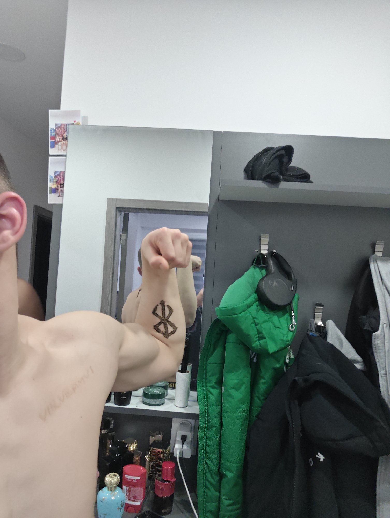 A person poses in front of a mirror, flexing their left arm to display a tattoo. The tattoo is a dark-colored symbol. A light-skinned person's upper body is visible, including part of their head and ear. Other details include toiletries on a shelf below the mirror, hanging coats, and headphones on a hook. Based on the text on the person's back, the date "VIII.VIII.MMVI" is apparent. The location is not discernible from the image provided.
