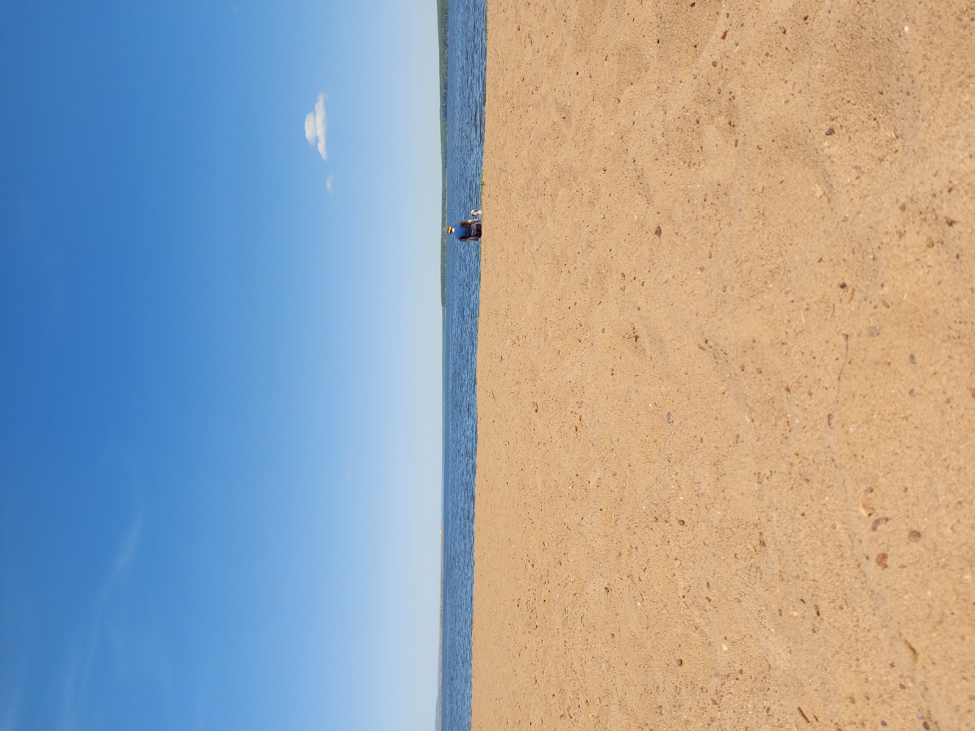 A vertically oriented photograph presents a scene composed of approximately one-third blue sky with a small cloud, one-sixth water with faint ripples, and one-half sandy shore. On the water's edge, a single person is visible, kneeling or squatting. The location is identified as Chaikovsky, Russia. Details about the person, the specifics of their actions, or any interactions are difficult to discern due to the distance. The environment appears to be a shoreline on a clear day.