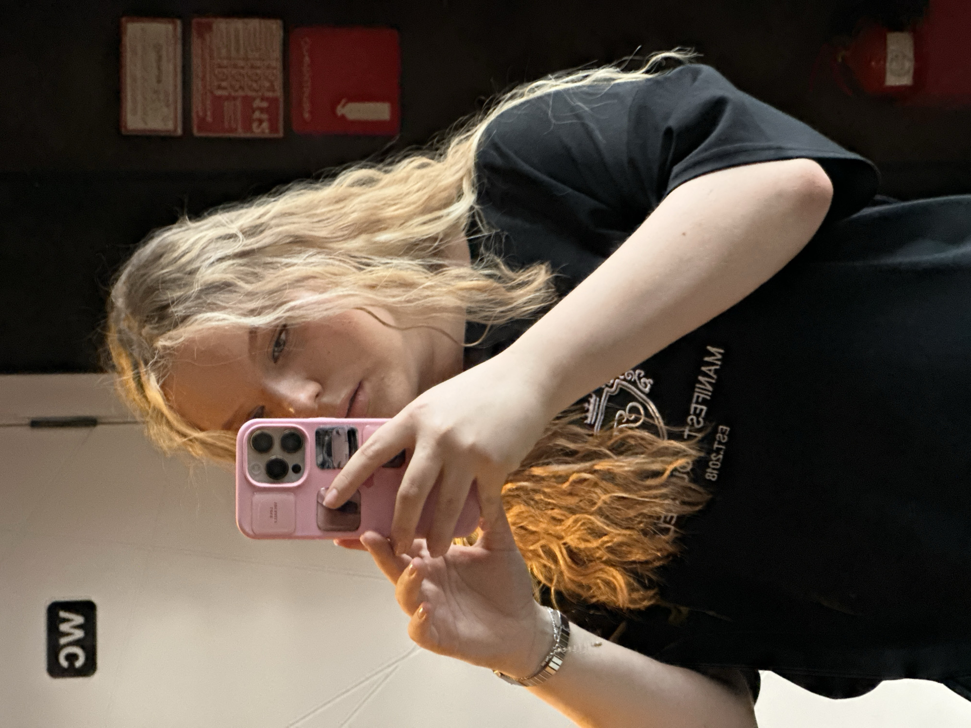 A young woman with blonde, wavy hair holds a pink iPhone and takes a selfie in a restroom mirror. She wears a black t-shirt with white lettering. The restroom has a white wall with a sign reading "MC" in black letters. Above the woman's head are three wall-mounted objects, two red and one light colored. A red fire extinguisher is visible in the background. The picture's location is the city of Chaikovsky, Russia.