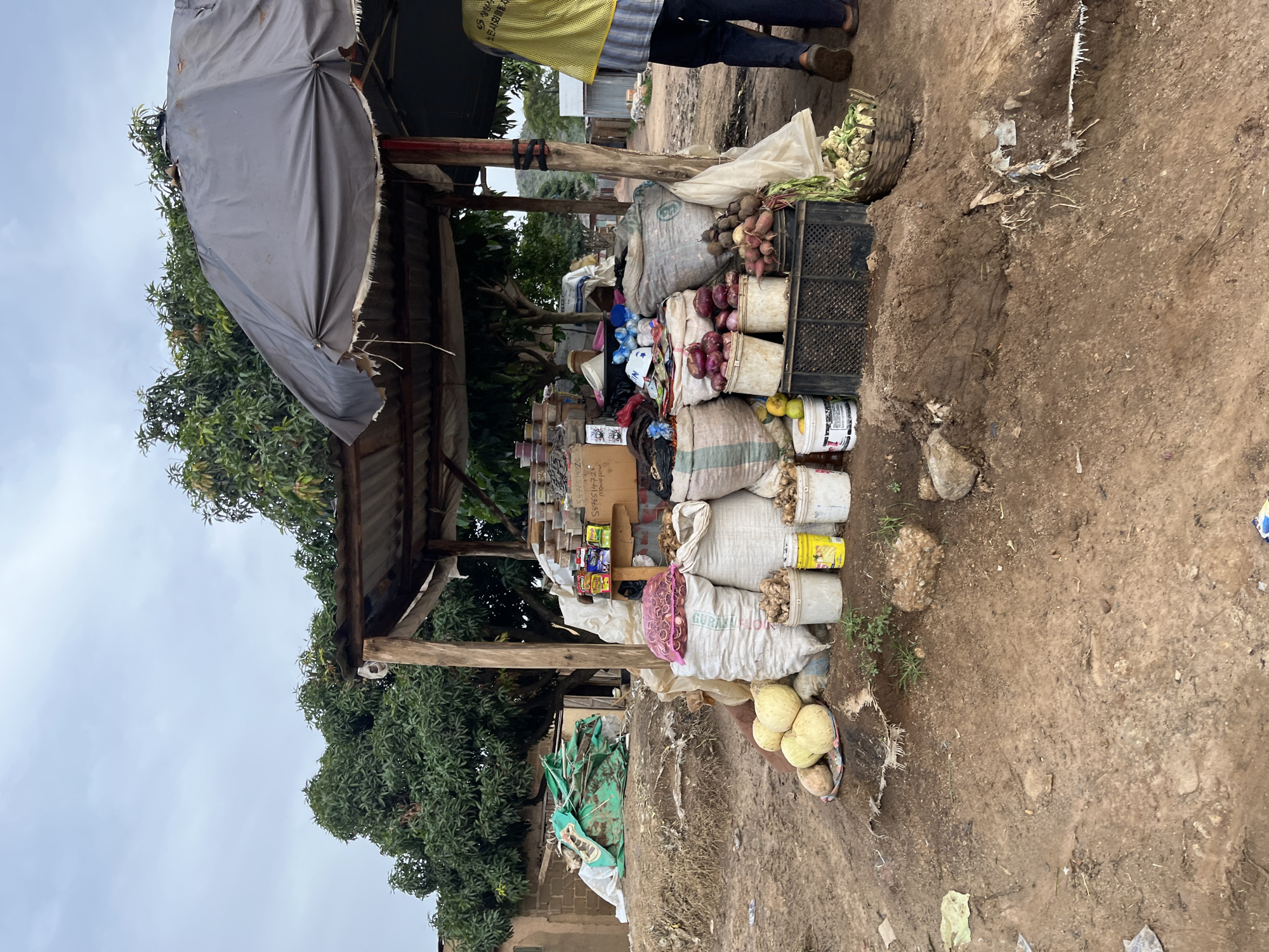The image depicts an outdoor market stall in Gura Top, Nigeria, on an overcast day. The stall is sparsely stocked with produce and other goods. A person, wearing a yellow vest over a striped shirt and dark pants, stands partially visible behind the stall. The stall itself is a makeshift structure with a corrugated metal roof supported by wooden posts. A grey tarp is draped over part of the roof.
Various items are displayed for sale, including large sacks of produce, some of which appear to be onions and root vegetables. Several white buckets contain what look like onions or garlic. In the foreground, a basket holds several pale yellow, round fruits, possibly gourds or melons, along with some smaller brown root vegetables. Smaller items, including packaged goods and boxes, are stacked on shelves within the stall. A green tarp is also visible, folded and placed near the stall. The ground is dirt, with some scattered debris.
The scene appears to be a typical market day, with goods laid out for potential customers. There are no strong emotional cues visible, and the overall impression is one of a functional, everyday commerce setting. No specific time of day can be discerned, other than it is daylight. The weather appears to be cloudy. A partially visible text on one of the sacks reads "GURAM".