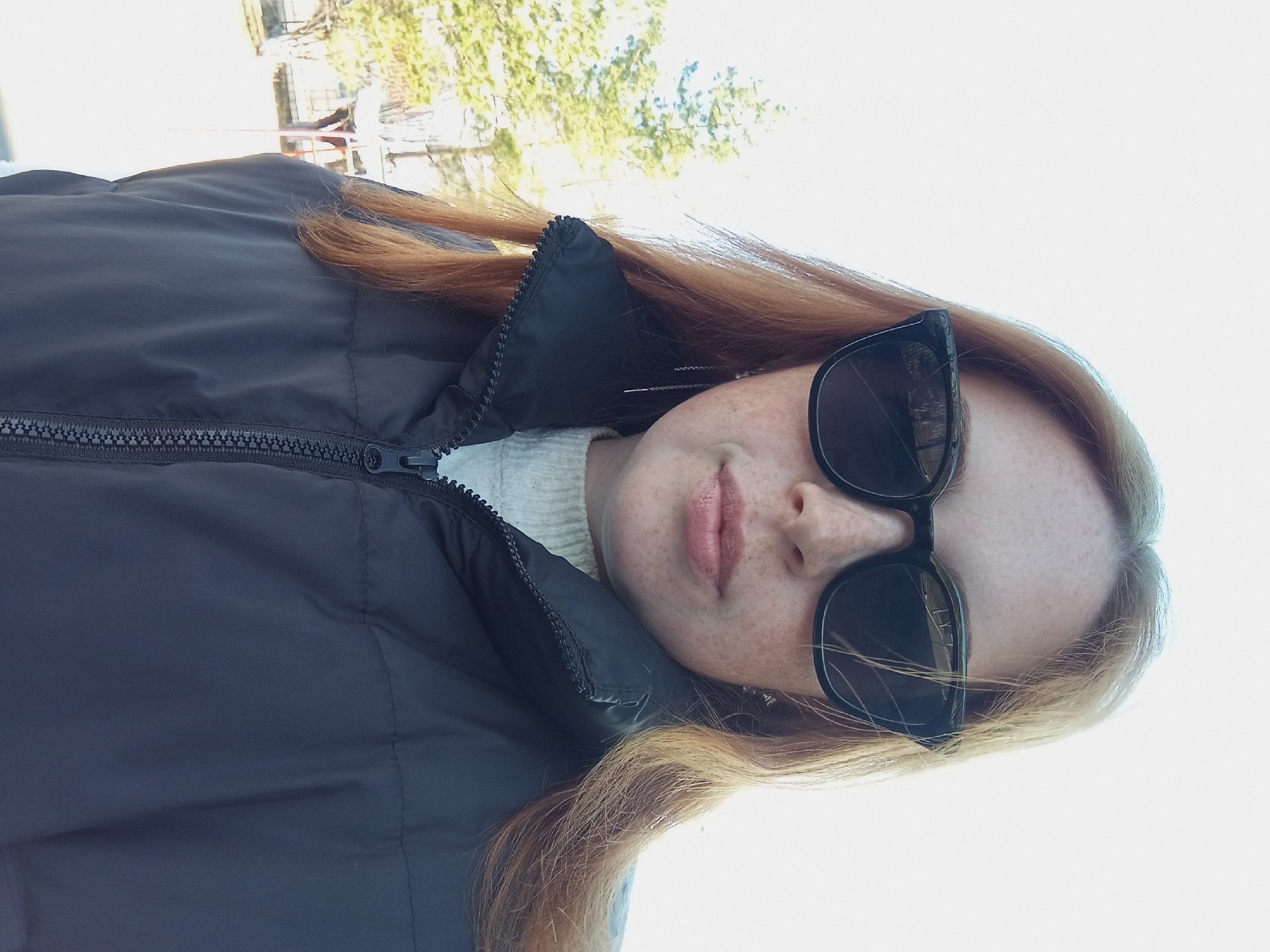 A woman with red hair and freckles is wearing sunglasses and a black puffer jacket with a white sweater underneath. The photo appears to be taken outdoors in Yurgamysh, Russia, indicated by the location provided, with trees visible in the background. The woman is looking at the camera.