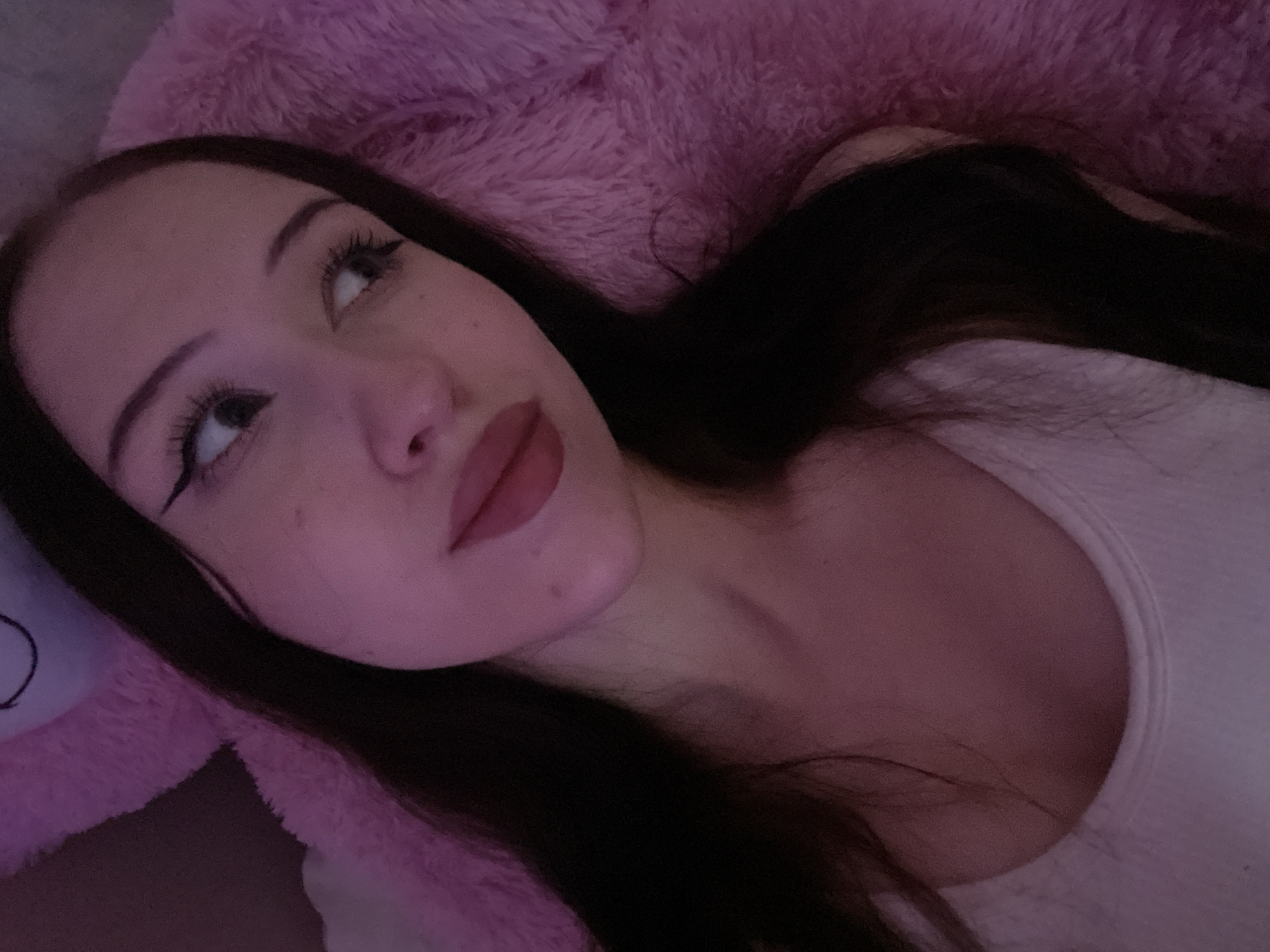The image shows a close-up, waist-up view of a young woman with long, dark hair, lying on a fluffy pink surface, possibly a blanket or stuffed animal. She is wearing a white tank top. Her face is turned to the right, and she is looking away from the camera. She has on dark eyeliner and lipstick. The lighting in the image is dim and tinted pink, suggesting an indoor setting with artificial light. There is no indication of the city of Votkinsk, Russia, or any specific location within it. The scene appears to be a personal, candid moment.