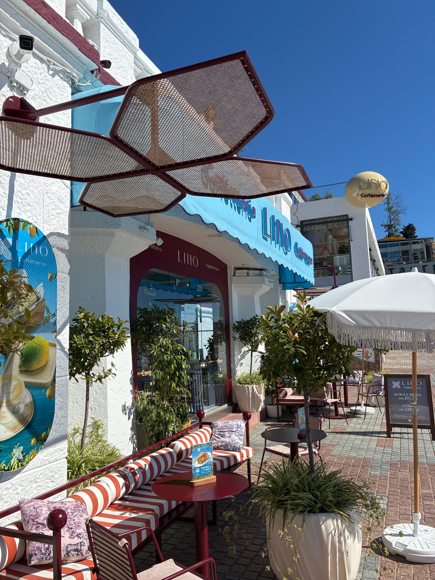 The image shows the exterior of a cafe called "Lino Caffetteria" on a sunny day. The cafe has a white facade with maroon accents. A blue awning with the cafe's name in red letters is extended over the entrance. There are outdoor seating areas with tables and chairs, some with red and white striped cushions. Several potted plants are arranged around the seating area. A white umbrella with fringe is open over one of the tables. A sign in Russian for "Lino Caffetteria" is visible. The cafe appears to be located in a city with cobblestone paving. The weather is clear and sunny with a bright blue sky.