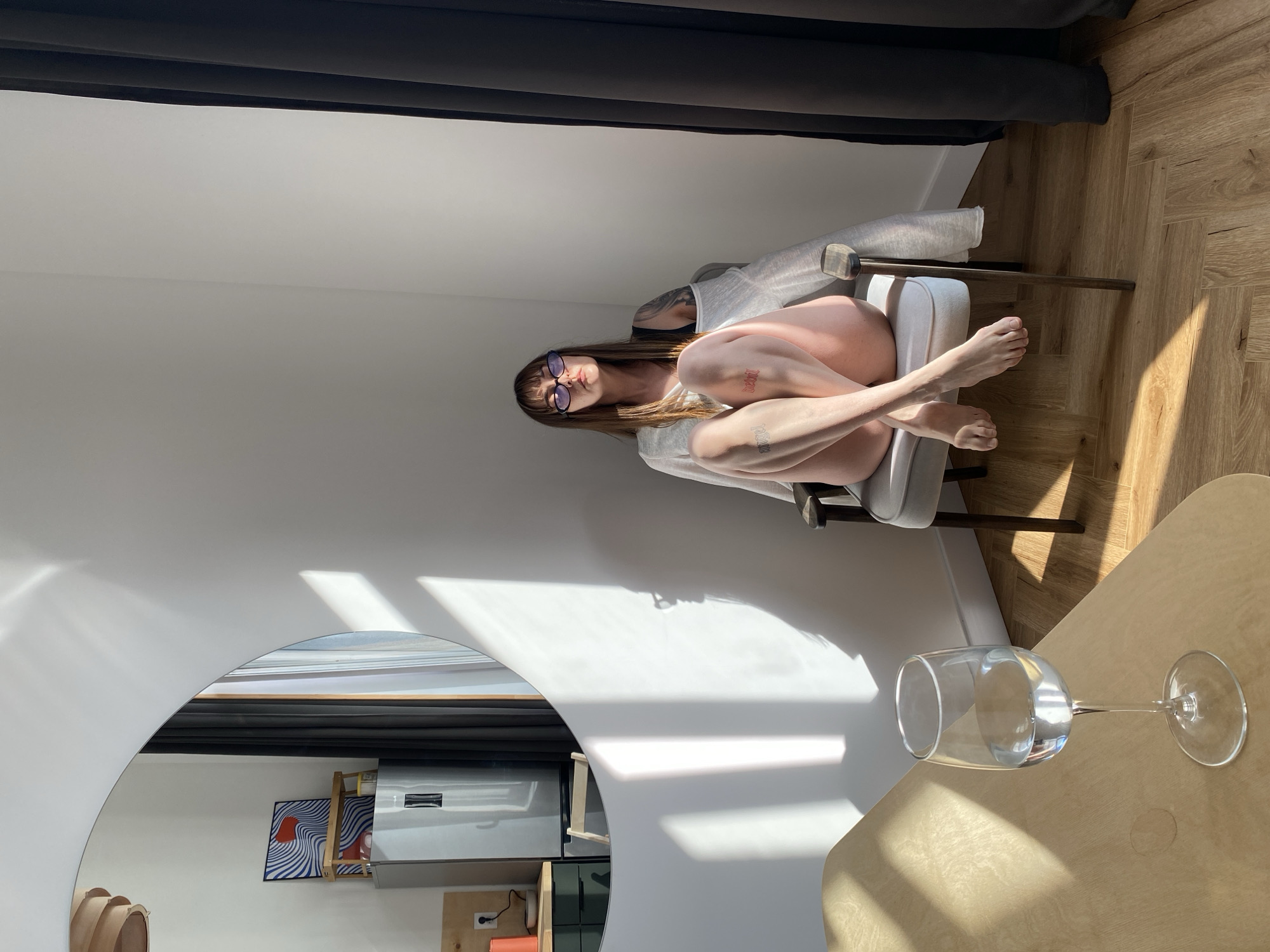 A woman wearing glasses sits in a chair in a sunlit room with hardwood floors. A table with a glass of water is in the foreground, and a mirror reflects a refrigerator and artwork on the wall. The location is a room in a building in Izhevsk, Russia.