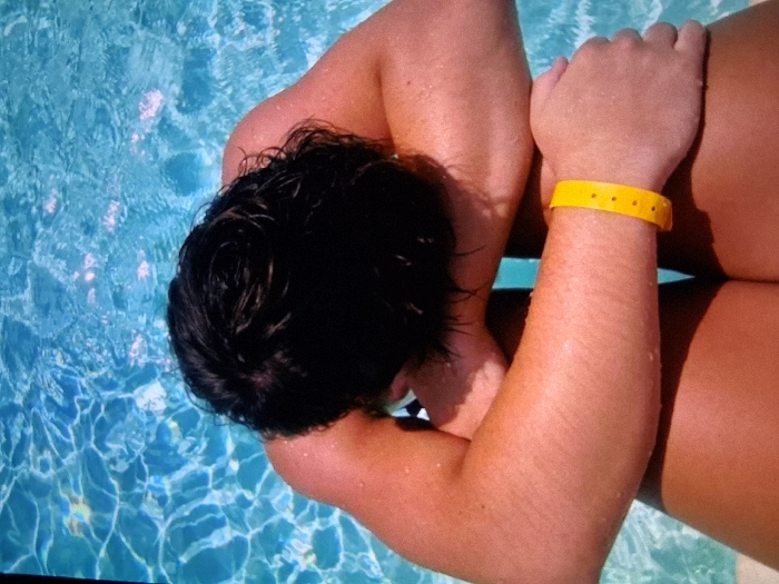An overhead image depicts an individual partially submerged in a light blue swimming pool. The subject's head is tilted forward, obscuring the face, revealing dark, wet hair. Both arms are crossed, resting upon the upper thighs. The skin on the visible arms and legs exhibits a tanned complexion, with numerous water droplets present. A bright yellow wristband, featuring five visible circular perforations, is worn on the right wrist. A dark garment is faintly visible near the neck/shoulder line and between the thighs. The pool water displays ripple patterns and reflected light. The scene is located in the city of Unknown, Unknown.