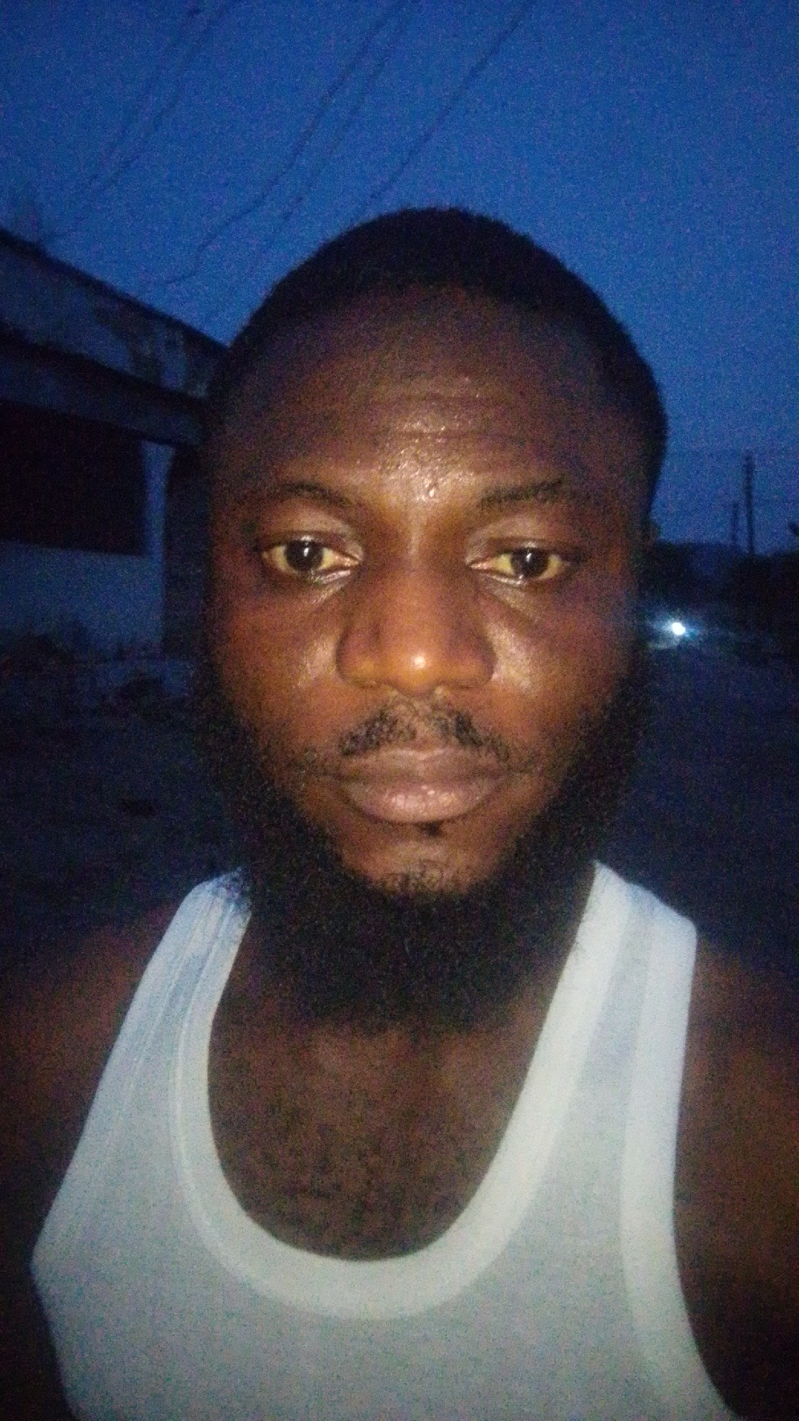 A close-up of a man wearing a white tank top. He has dark skin, a beard, and short black hair. His face appears slightly shiny, possibly due to sweat. The background is dark, with what seems to be the corner of a building and utility lines visible in the upper portion of the image. A possible source of light appears blurred at a far distance on what seems to be a road. No specific details confirm the location as Lokoja, Nigeria, and the image only offers a general outdoor night setting.
