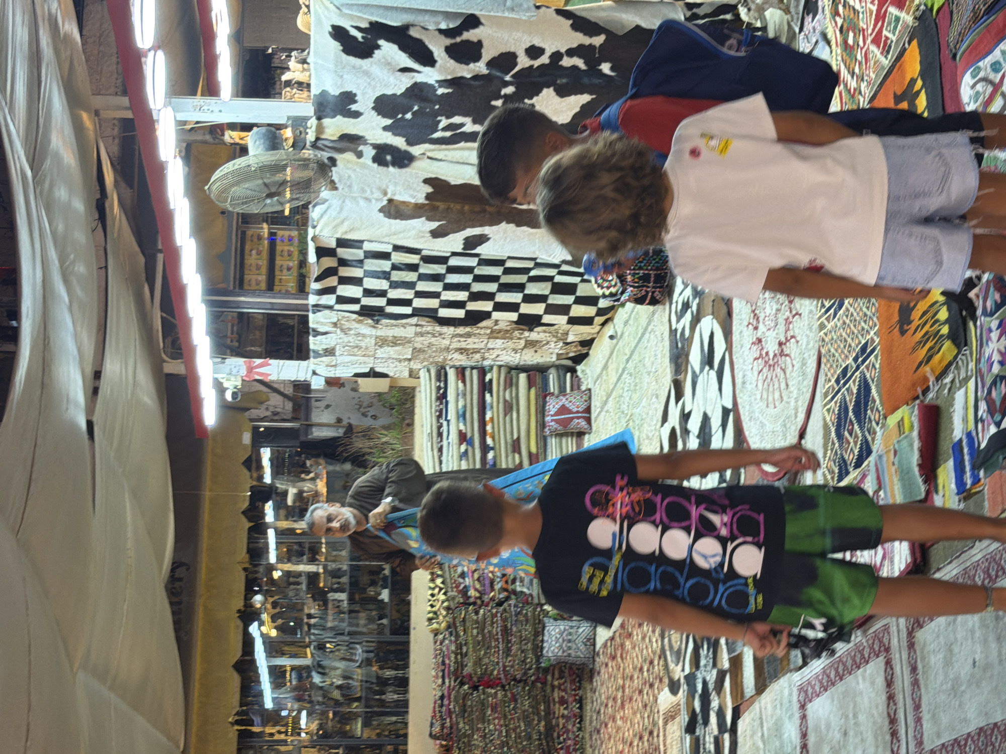 The image depicts a busy market scene in Sharm El Sheikh, Egypt. In the foreground, two young boys are viewed from behind, one wearing a black t-shirt with a graphic print and green shorts, and the other in a white t-shirt and denim shorts. They appear to be looking at a man in the middle ground. This man, who has a grey beard and is wearing a dark brown robe, is holding up a vibrantly colored textile, possibly a rug or tapestry. Behind him, stacks of textiles, likely rugs and carpets, are displayed on shelves and on the ground, creating a visually rich environment. Further in the background, shelves filled with various items, possibly souvenirs or traditional crafts, are visible behind a glass display. Overhead, a white canopy is stretched across the market, with lighting fixtures illuminating the space. A large fan is also visible in the background. The overall impression is of a lively and bustling marketplace, characteristic of tourist areas in Sharm El Sheikh.