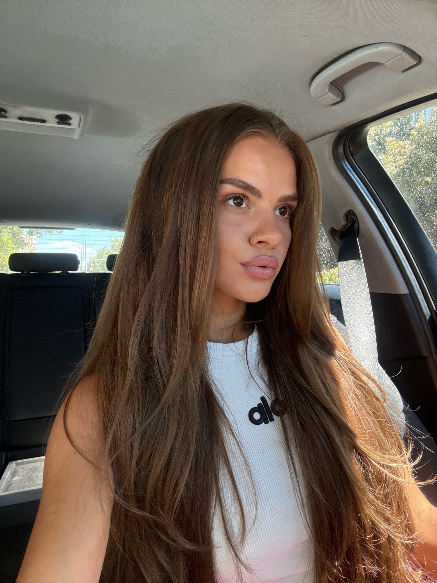 A young woman with long, brown hair is sitting in the passenger seat of a car. She is wearing a white ribbed tank top with a black logo that reads "alo". The car appears to be parked on a street in Plovdiv, Bulgaria, with trees and buildings visible through the windows. The sun is shining, casting shadows on the woman's face and hair.