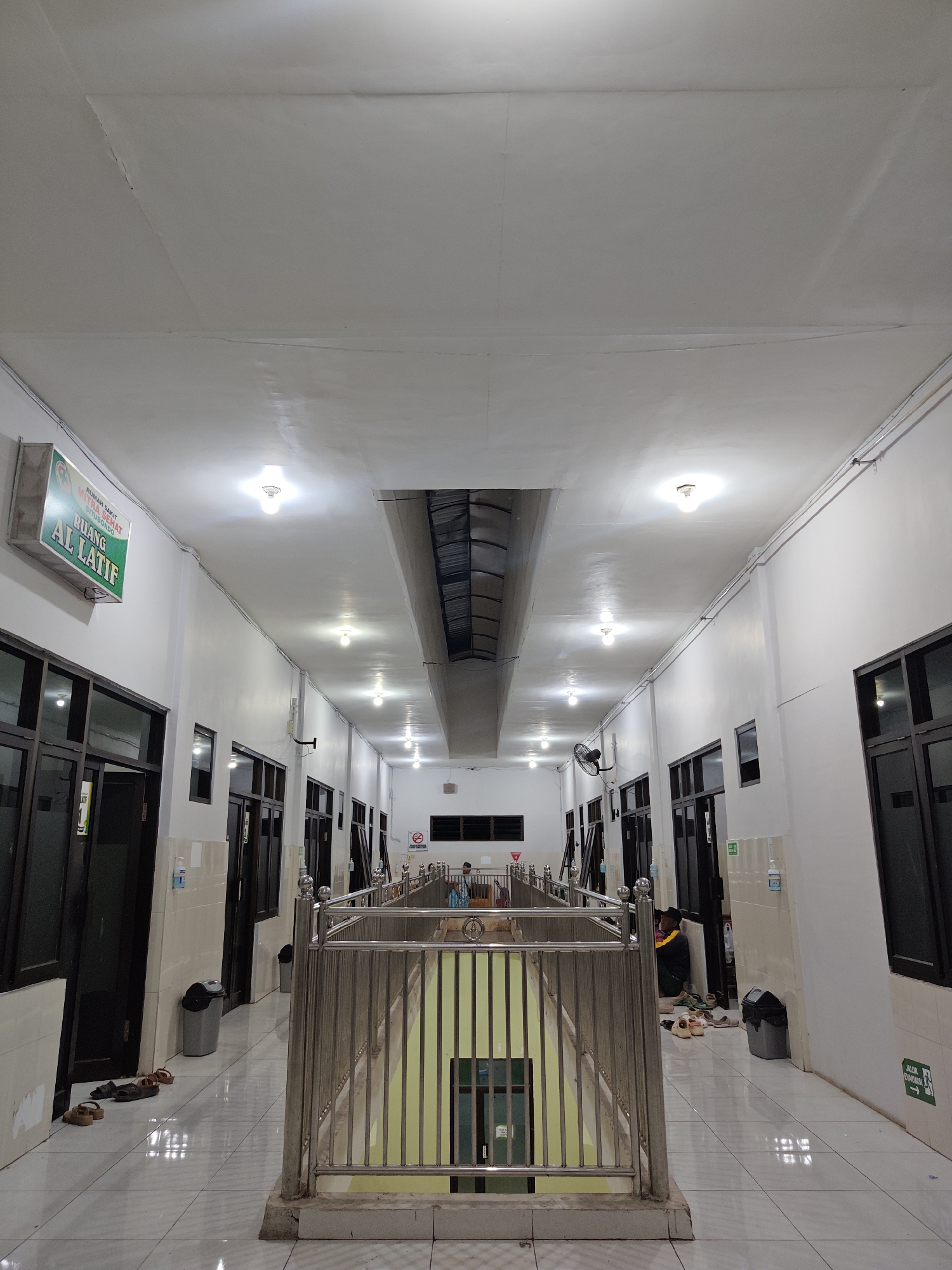 The image depicts a long, brightly lit corridor within what appears to be a hospital or similar institution. A sign on the left wall reads "RUMAH SAKIT WIJAYA SEHAT SITUBONDO RUANG AL LATIF," indicating it is the Al Latif room in Wijaya Sehat Hospital in Situbondo, Indonesia. The corridor is lined with doors on both sides, most of which are closed and made of dark wood with glass panels. The walls are painted white, and the ceiling is also white with several overhead light fixtures.
In the foreground, a metal railing encloses a central area that appears to be a stairwell or a lower level, filled with natural light. Several pairs of sandals are scattered on the tiled floor along the corridor. Further down the corridor, a few individuals are visible. On the left, near a doorway, a person is sitting on the floor. On the right, in the distance, two people are seated, one appearing to be a healthcare professional in uniform. A fan is mounted on the wall on the left side of the corridor. Trash cans are placed strategically along the walls. An "Emergency Exit" sign with an arrow pointing right is visible on the right wall. The overall environment suggests a functional, sterile, and somewhat institutional setting, likely a waiting area or a passage within a medical facility.