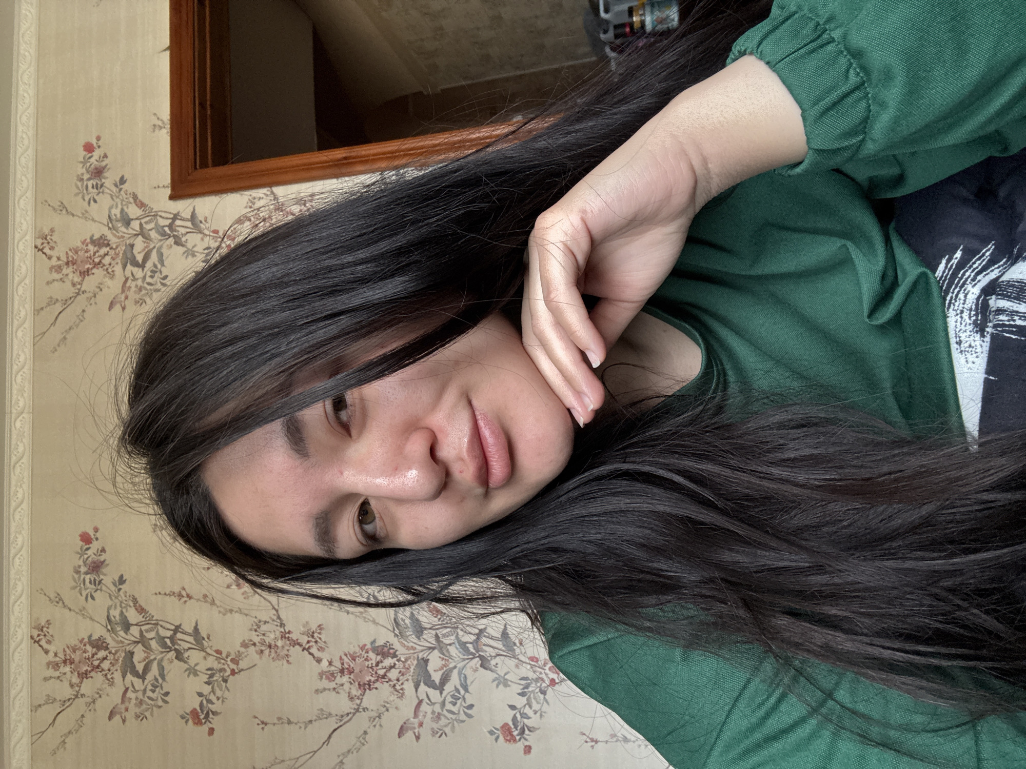 A person with long, dark hair is seen in a close-up selfie. Their face is angled towards the camera with their hand resting on their cheek. They are wearing a green shirt. The background features floral-patterned wallpaper and a wooden-framed object, possibly a mirror or picture frame. The presence of the patterned wallpaper and wooden frame suggests the image was taken indoors, possibly in a domestic setting. There is no verifiable information in the image or context clues suggesting the location is Kurovskoye, Russia, so I cannot provide any specific information about that location.