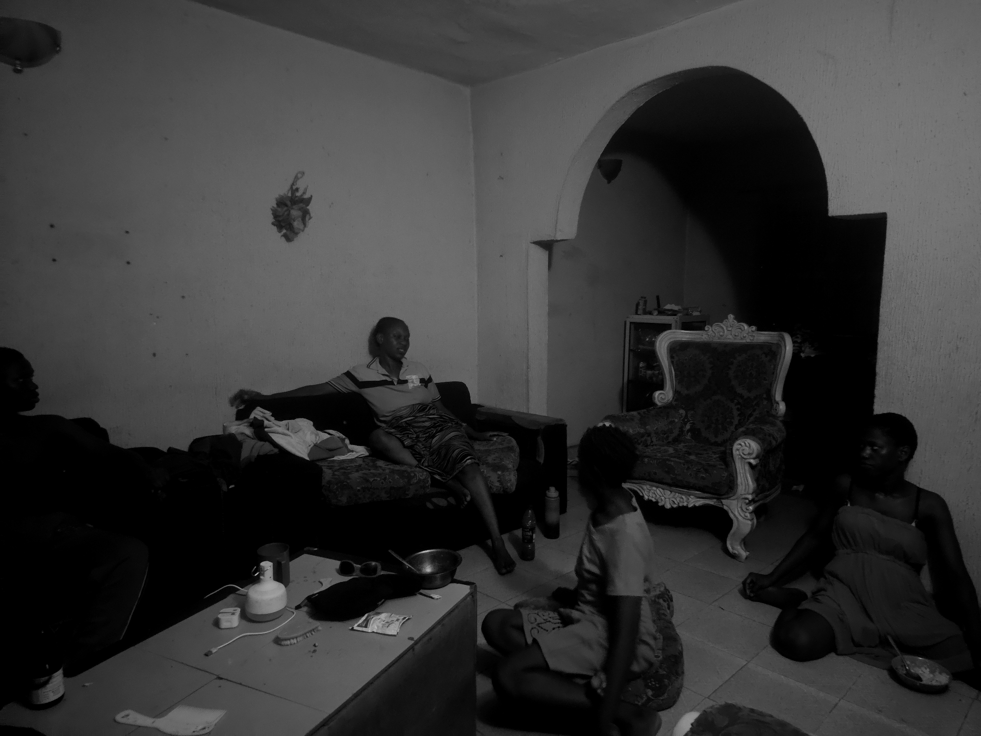 The black and white photograph captures an indoor scene, likely a living room, with several people gathered. At least five individuals are visible, appearing to be adults or young adults, mostly with dark skin tones. On the left, a person is partially visible, seated on a dark couch. Further along the same couch, a person wearing a striped polo shirt and a patterned skirt or sarong is seated with their legs outstretched, barefoot.
In the foreground, a low rectangular table holds various items including a white pump bottle, a phone charger, a USB cable, sunglasses, a metal bowl, a brush, and a small white plastic scoop or spatula. To the right of the table, a person in a light-colored top and patterned bottom is seated on the tiled floor, facing slightly away from the camera. Further right, another person in a dark dress or top and skirt is seated on the floor, next to an ornate armchair with a high back and carved details. A small bowl containing food or liquid is placed near this person.
The room features light-colored, textured walls. An arched doorway on the right leads to a darker interior space, where a shelf with items is faintly visible. The lighting is dim throughout the room, suggesting it might be evening or an interior with limited light sources. The atmosphere appears casual and relaxed, with people engaged in either conversation or simply resting. No visible text is discernible in the image.