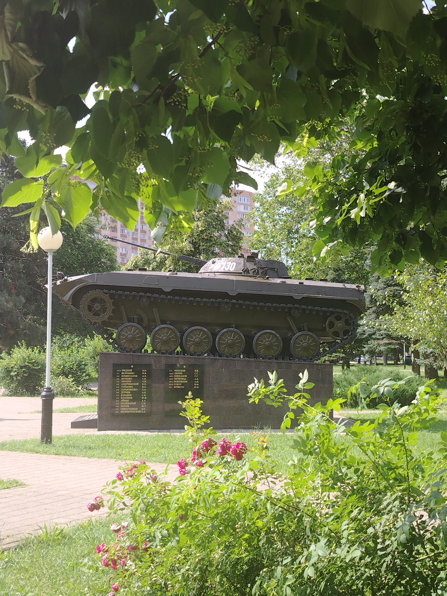 The image captures a memorial dedicated to soldiers, featuring a Soviet-era infantry fighting vehicle, likely a BMP-1, mounted on a granite pedestal. The vehicle is painted in a dark green camouflage and has the number "2130" visible on its turret. The pedestal displays names of soldiers, indicating a military memorial. The scene is set outdoors in a park-like environment, with lush green trees and bushes, including a flowering rose bush in the foreground. A lamppost is visible to the left of the tank. In the background, a multi-story residential building can be seen, suggesting an urban setting. The lighting indicates a sunny day. This memorial is located in the city of Krasnodar, Russia, specifically in the city park named after the 70th anniversary of the Victory in the Great Patriotic War, on the territory of the military commissariat of Krasnodar.