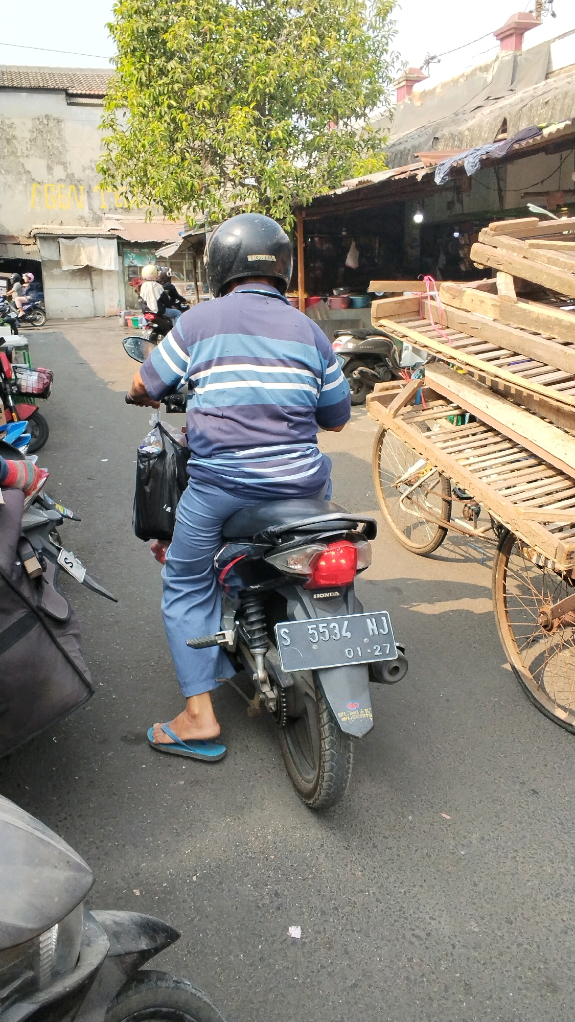 An adult male wearing a blue and white striped polo shirt, blue pants, and a black helmet rides a black motorcycle in a street scene that is likely in Mojokerto, Indonesia. The man is seated on the motorcycle, facing away from the camera. He has a black plastic bag hanging from the handlebars, and his right foot is resting on a blue flip-flop. The motorcycle's license plate reads "S 5534 NJ 01-27".
To the right of the motorcycle, a wooden cart with two wheels is visible, piled high with wooden planks and other materials. The cart is weathered and appears to be old. In the background, there are buildings with corrugated metal roofs and trees with green foliage. Other motorcycles and a glimpse of a person on a scooter can be seen in the distance. The ground is paved with asphalt. The scene suggests a busy street or market area.
