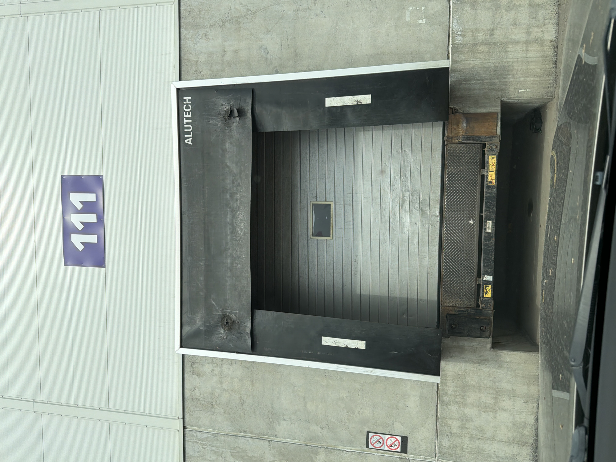 The image shows a loading dock with the number "111" on the adjacent wall. The loading dock door has the word "ALUTECH" printed vertically on its side. The loading dock is surrounded by concrete walls. The location is a building in Sluchainyi, Russia.