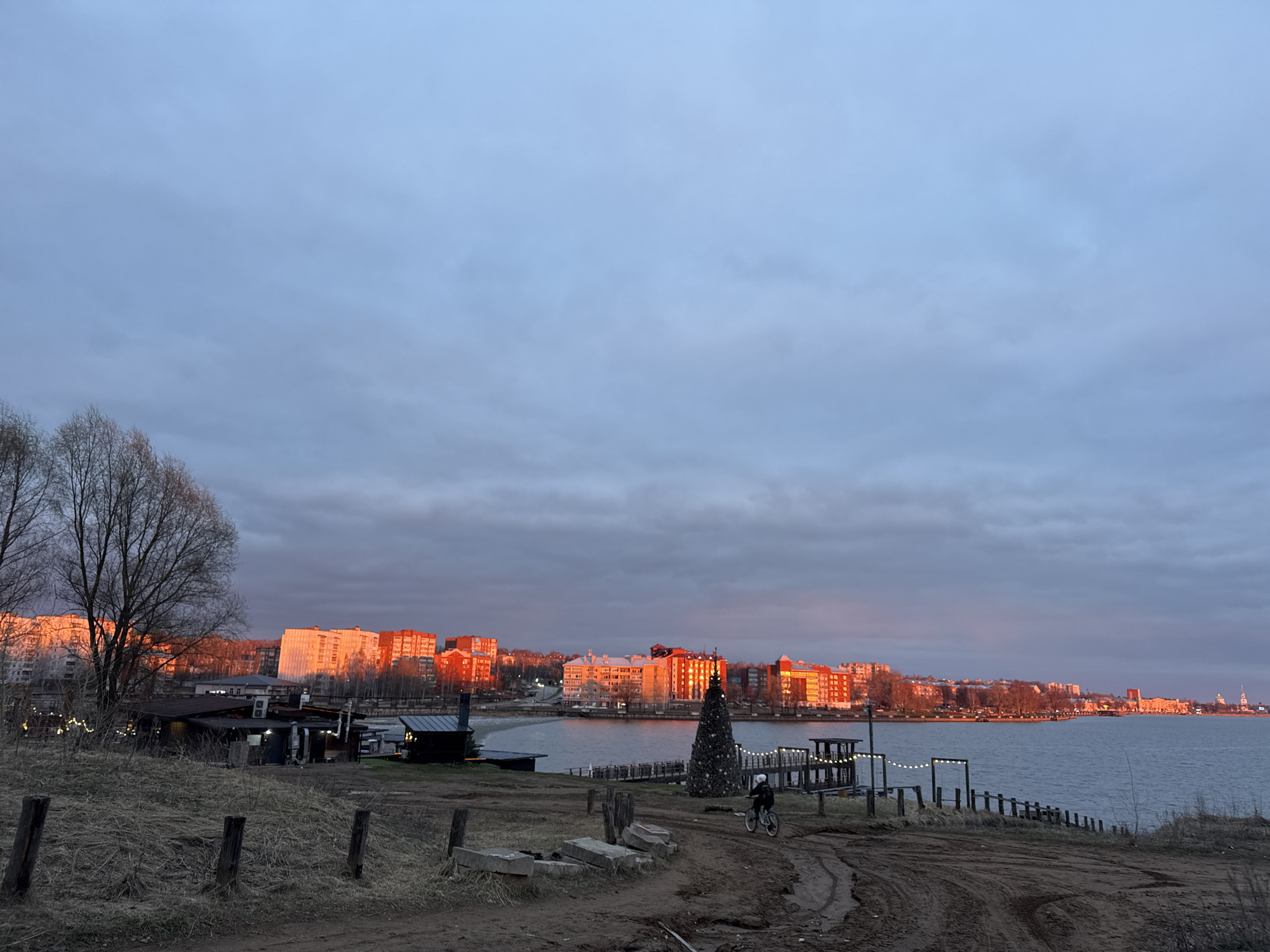 This panoramic image captures a tranquil scene along the Votkinsk Reservoir in Votkinsk, Russia, during either sunrise or sunset. The sky is overcast with shades of grey and muted blue, providing a soft, diffused light, yet the low-angle sun casts a striking warm, orange-red glow upon the facades of numerous multi-story apartment buildings and other structures lining the distant shore.
In the foreground, a muddy, unpaved dirt path winds through dry, sparse grass and scattered concrete blocks and wooden posts. Bare deciduous trees with thin branches stand to the left, and smaller, dark-roofed shacks or small buildings are visible near the water's edge. A notable detail is a large, conical, decorated structure resembling a Christmas tree, adorned with lights, positioned near the water in the mid-ground. Further along the shoreline, a string of festive lights hangs between wooden posts, illuminating a small pier or platform extending into the water.
The main activity in the scene is a single person, wearing a white helmet, riding a bicycle along the dirt path in the mid-ground. The body of water, likely the Votkinsk Reservoir, is calm and reflects the subdued light of the sky. The overall weather appears cool, possibly early spring or late autumn, indicated by the bare trees and the general atmosphere. No visible text is discernible in the image.