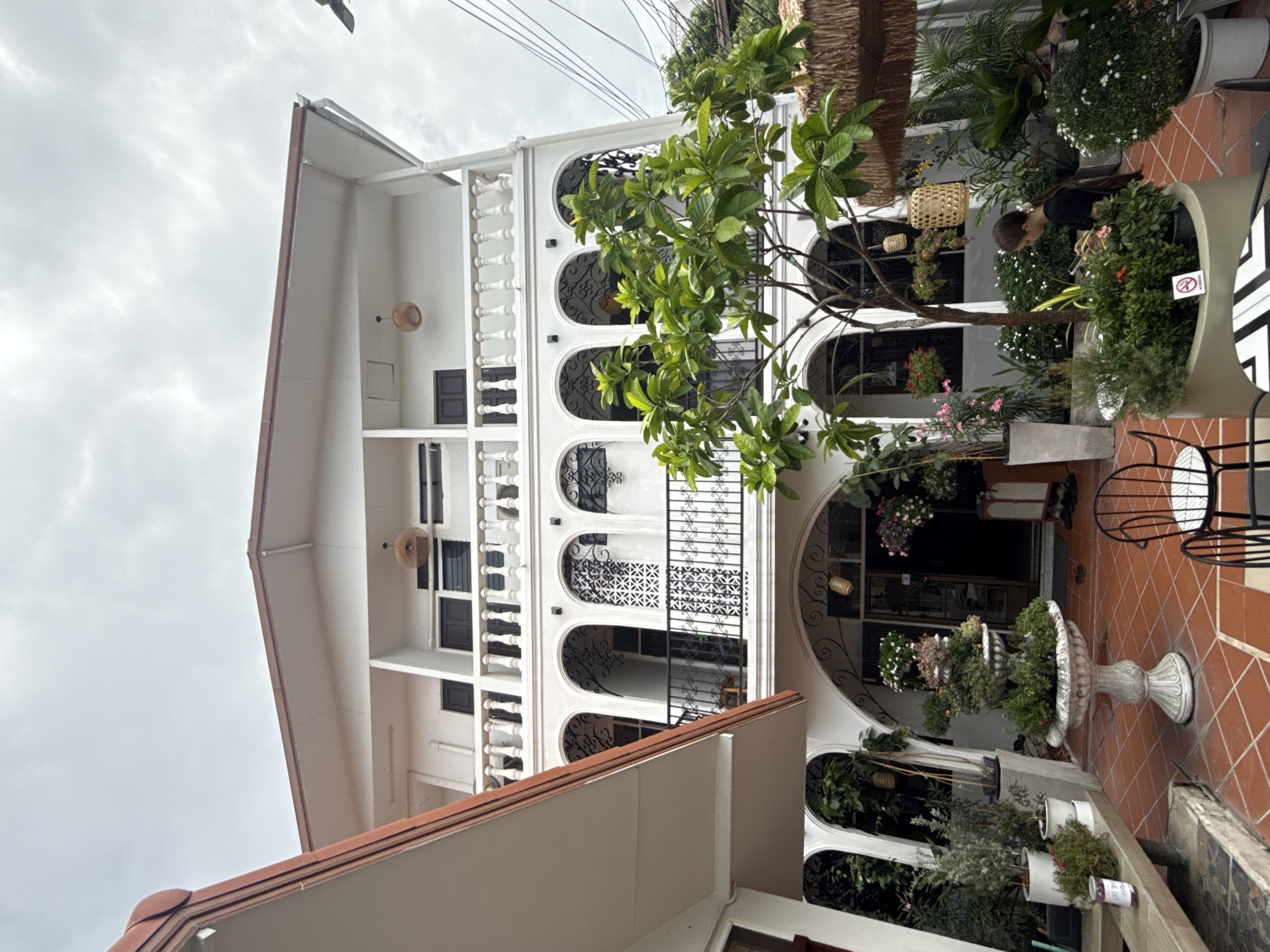 The image depicts the exterior of a multi-story building in Bangkok, Thailand, characterized by its white facade, arched windows with decorative metalwork, and balconies with white balustrades. The architectural style suggests a blend of colonial and tropical influences. In the foreground, a paved courtyard area is visible, with terra cotta tiles and an assortment of potted plants and trees, some of which are flowering. A large, ornate fountain stands centrally, and several outdoor chairs and tables are present, suggesting a cafe or restaurant setting. A woman in a black shirt is seen interacting with her phone amidst the greenery. The overall impression is of a tranquil, well-maintained outdoor space connected to a building, likely a commercial establishment. The sky is overcast, indicating potentially cloudy weather.