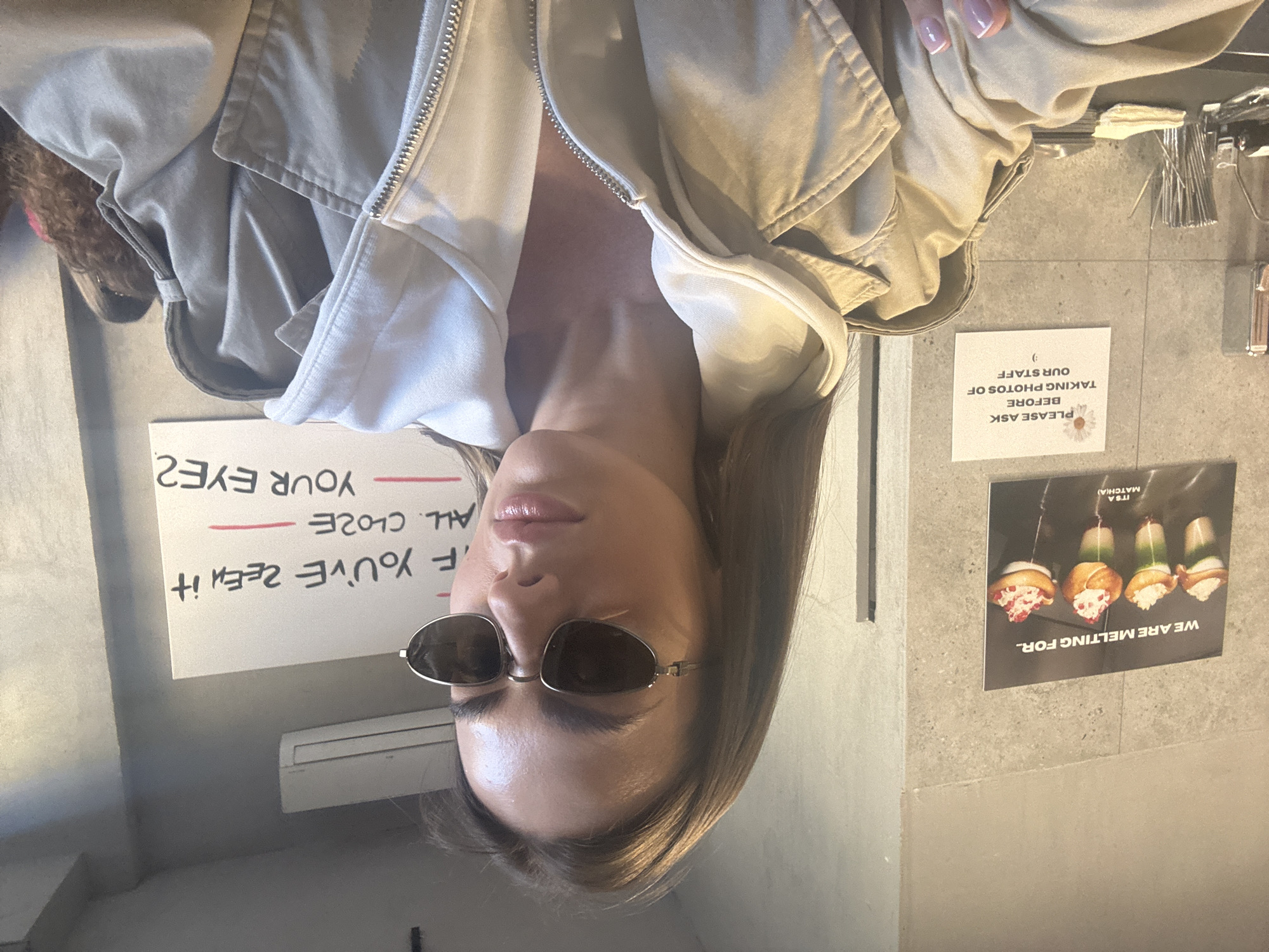 A woman wearing sunglasses and a light-colored jacket is indoors in Warsaw, Poland. Signs on the wall read "WE ARE MELTING FOR.. IT'S A MATCH(A)," "PLEASE ASK BEFORE TAKING PHOTOS OF OUR STAFF," and "IF YOU'VE ZEEN IT ALL CLOZE YOUR EYE2."